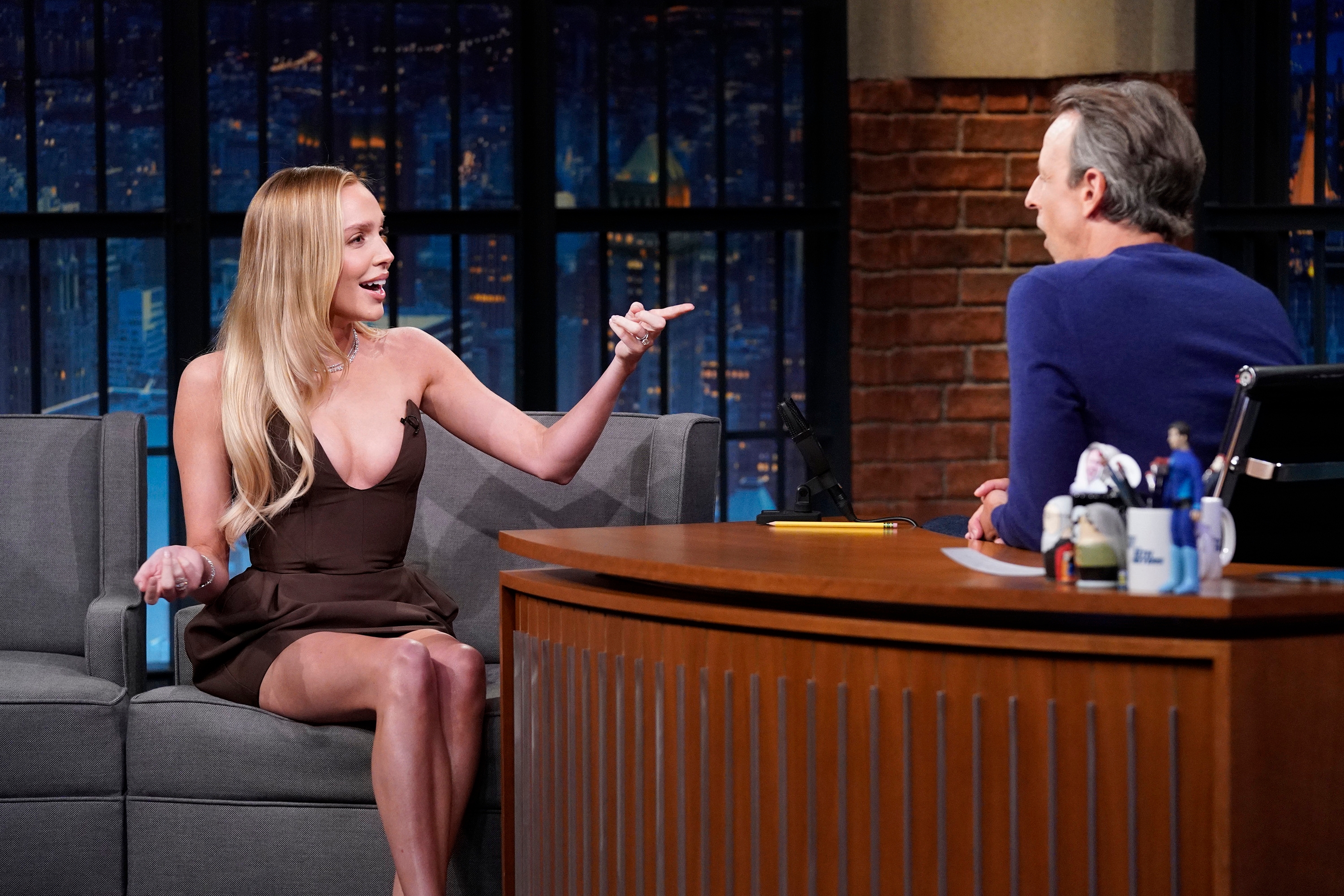 Alex Cooper in a plunging dress chats animatedly with Seth Meyers seated behind a desk decorated with figurines