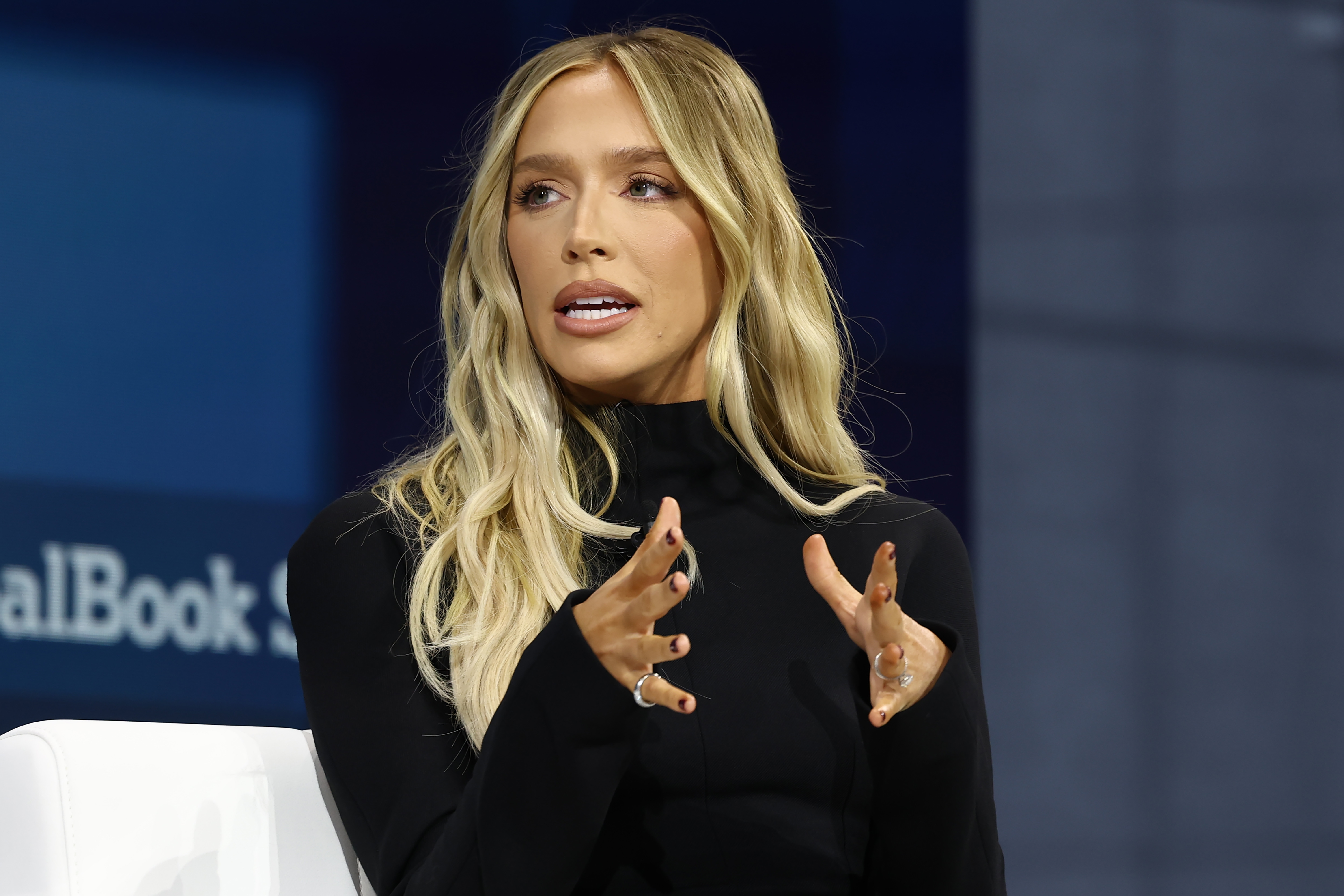 Alex Cooper with long hair gestures while speaking on stage, wearing a turtleneck outfit