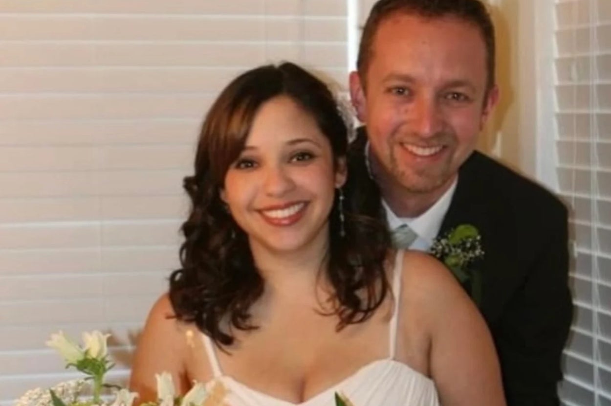 A smiling couple poses together indoors, wearing formal attire. She wears a sleeveless dress, and he is in a suit with a tie, holding a bouquet