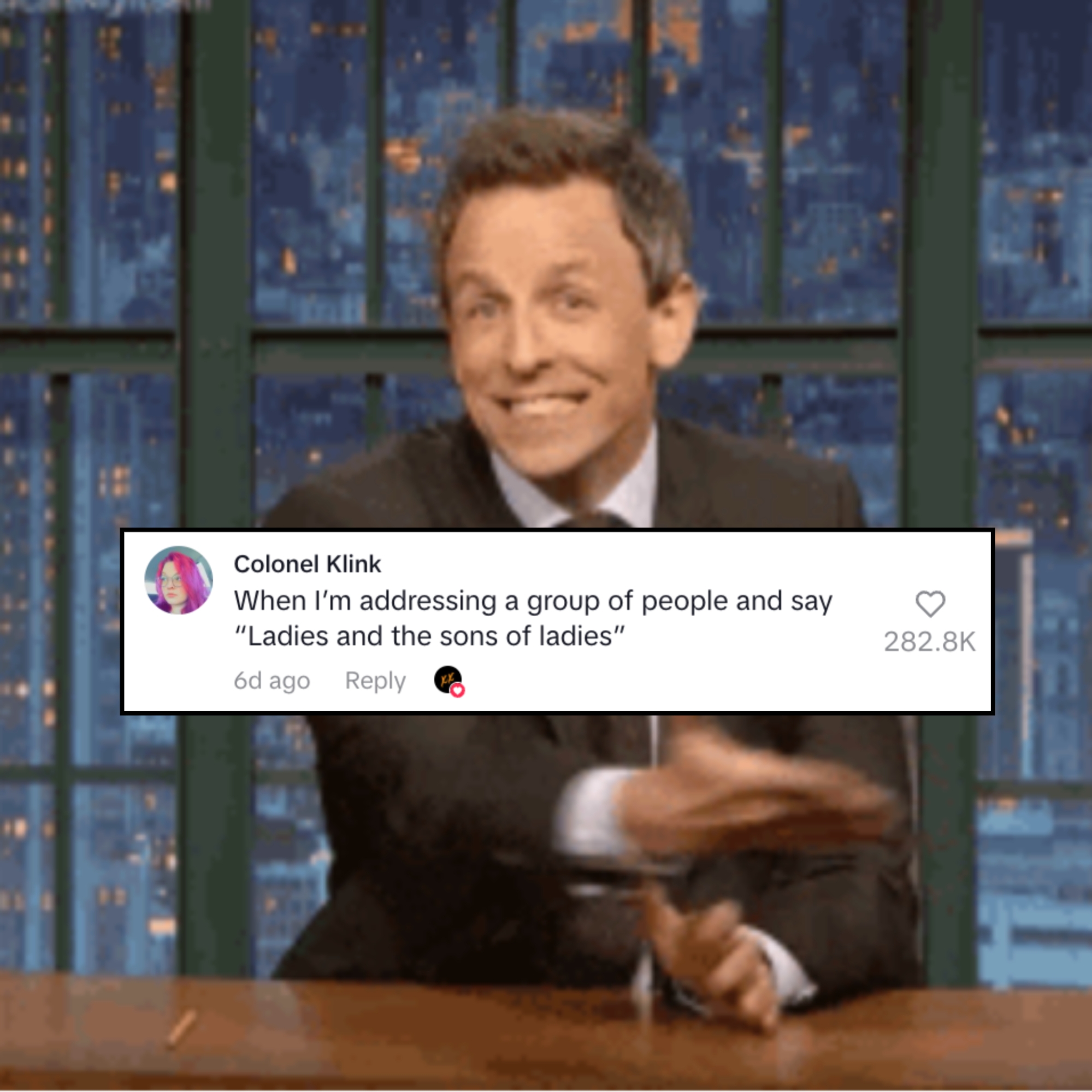 En person i dress sitter ved et skrivebord på et talkshow -sett, smiler og gestikulerer entusiastisk