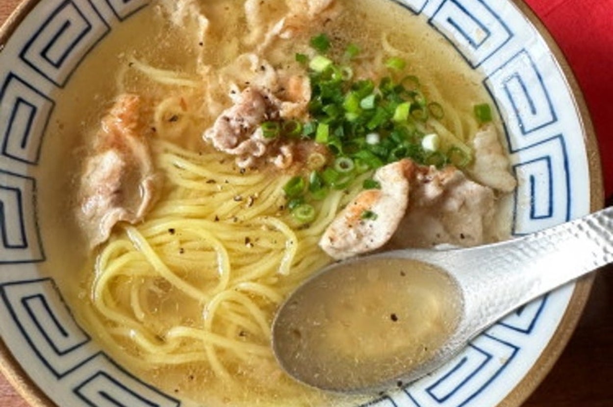 ラーメンの丼にスープ、麺、豚肉、ネギ。右にはスプーンが置かれ、横に赤い布が見える。