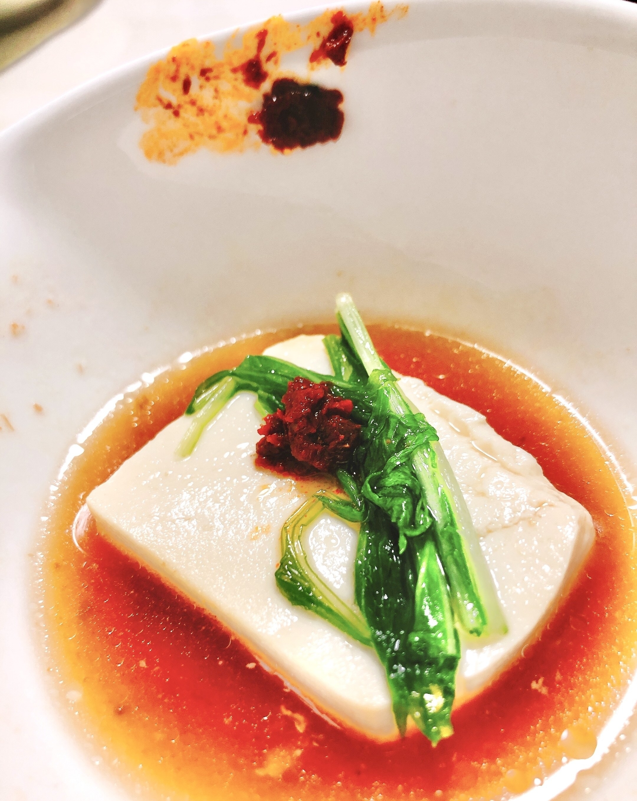豆腐の上に緑の野菜と赤い調味料が添えられた料理。ソースがかかった白い皿に盛られています。