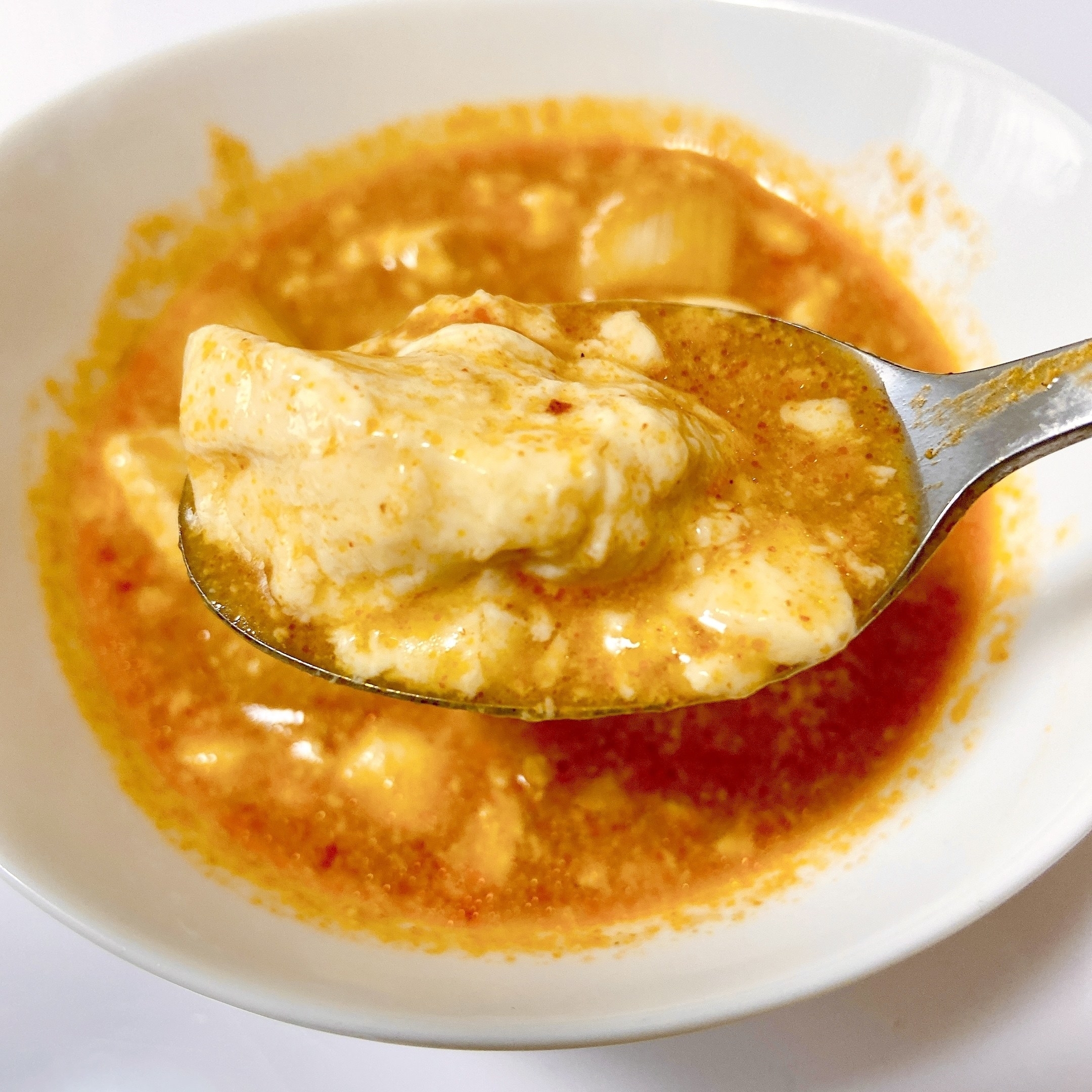 スプーンで持ち上げたマーボー豆腐が入った白いボウル。クリーミーでスパイシーな一品。