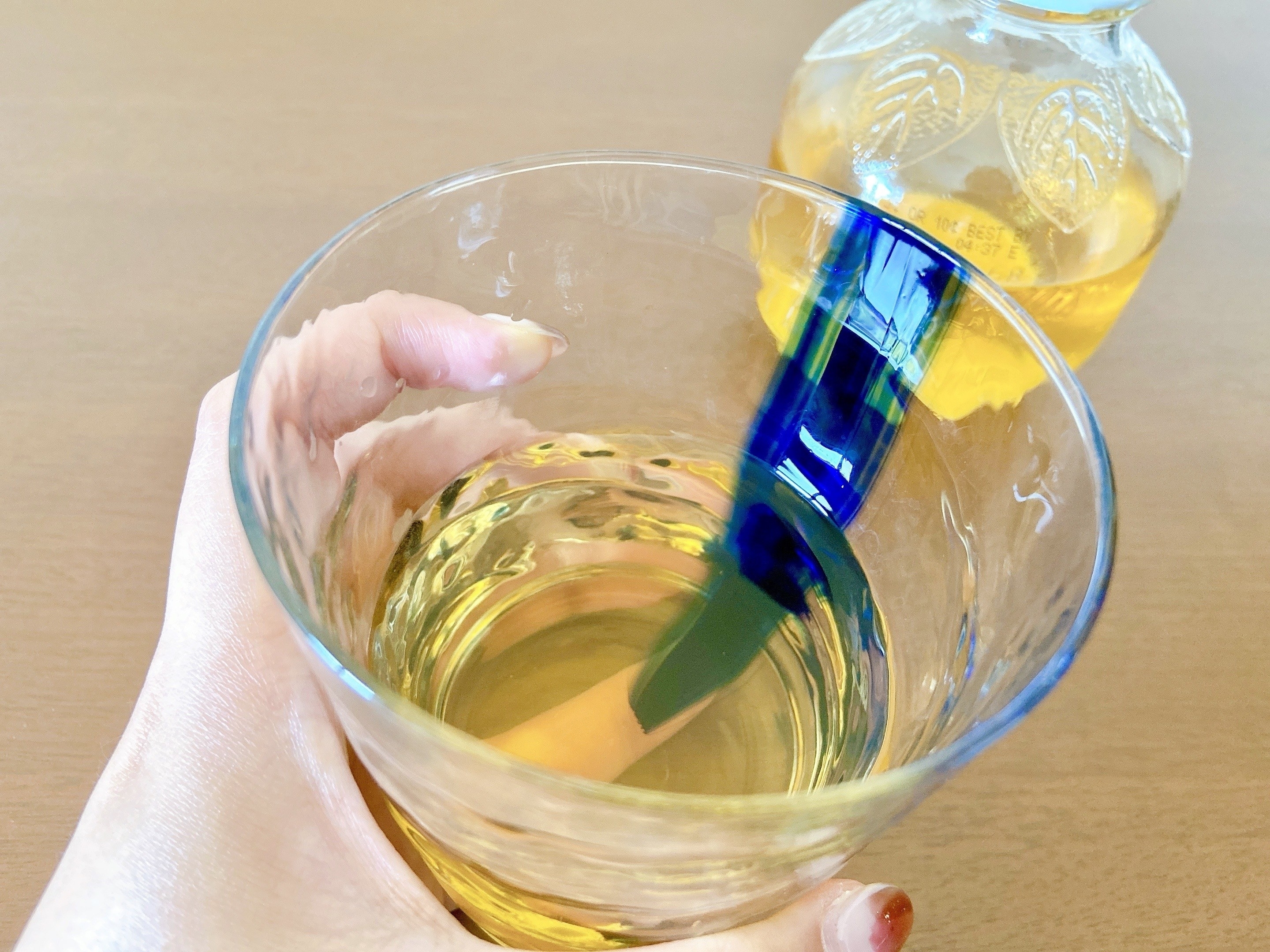 手に持ったグラスに黄色い液体が注がれており、背景に同じ液体のボトルが見える。