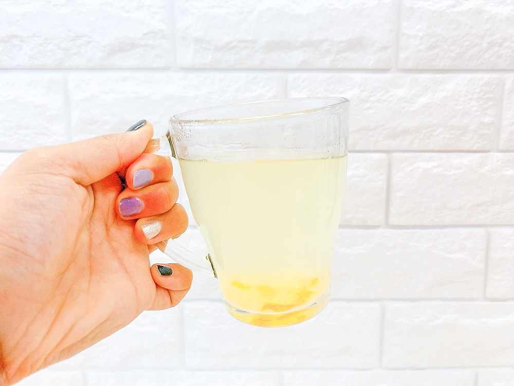 手に持ったガラスのカップに、透明の飲み物と果物のようなものが少し入っている。背景は白いレンガ模様の壁。