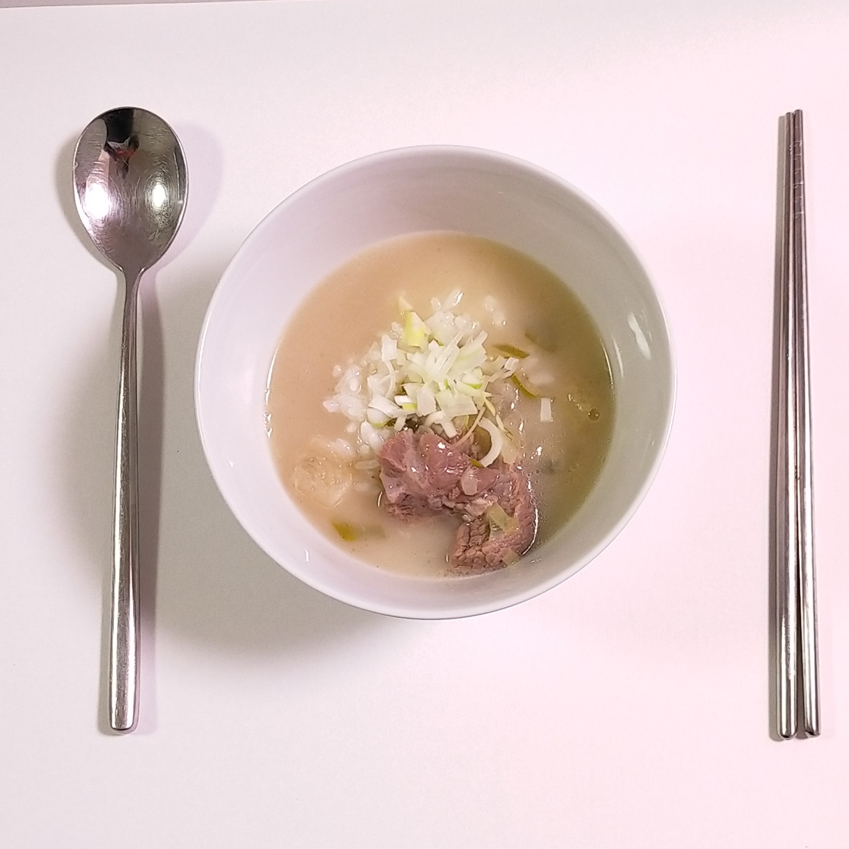 白いテーブルに底の深い白いボウル。中にはスープ、ネギと肉。フォークとスプーンが並ぶ。
