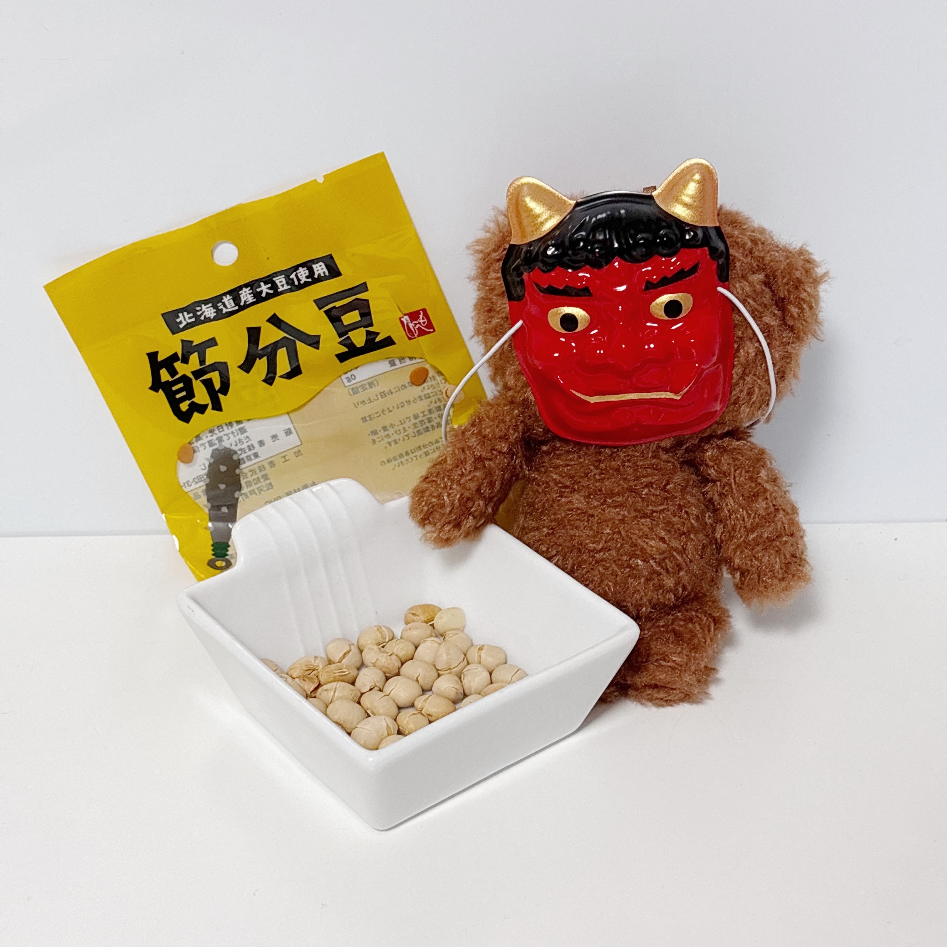 赤鬼のお面をかぶったぬいぐるみが豆のパッケージと皿のそばに座っています。節分を象徴する光景です。