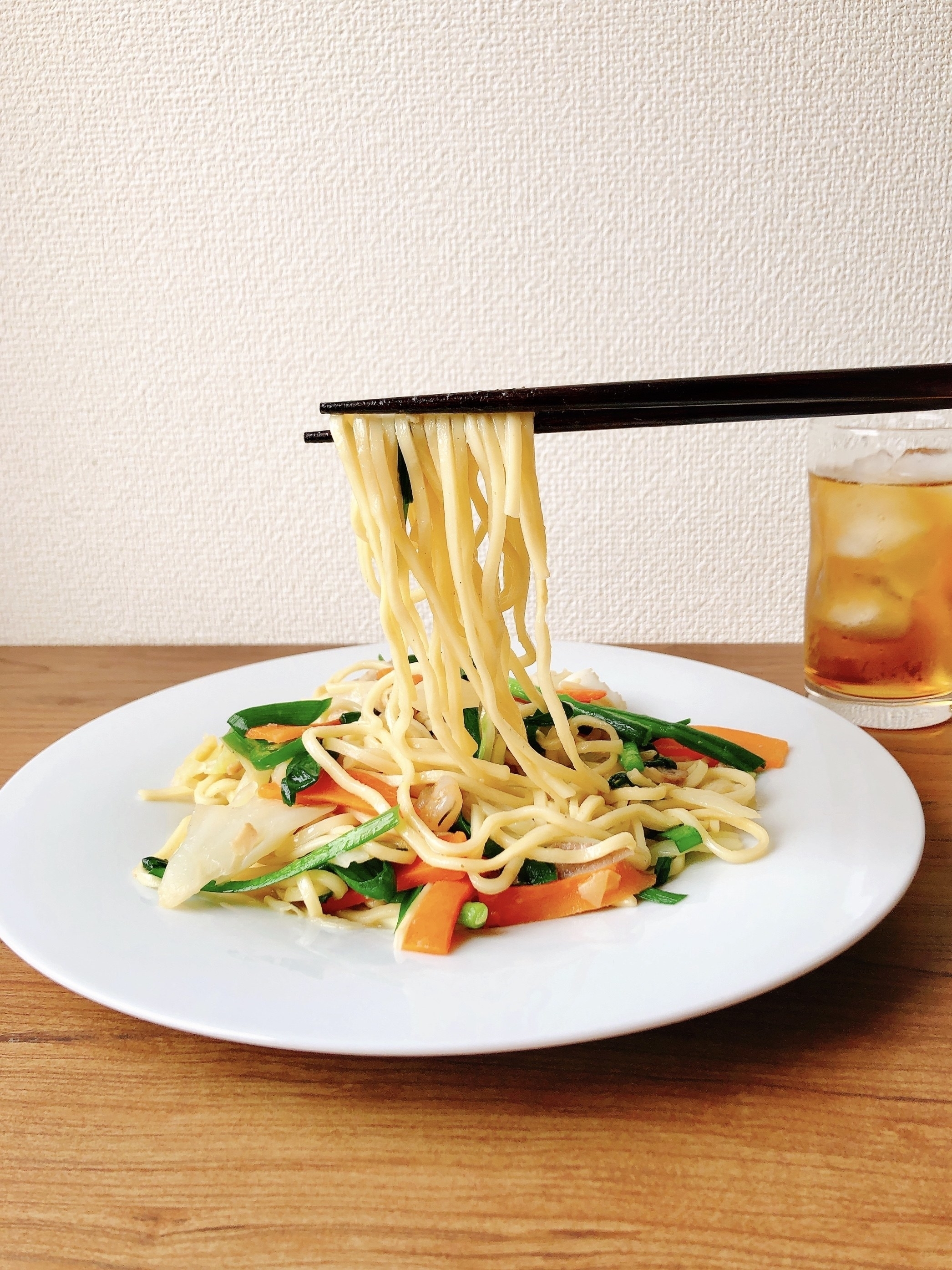 麺と野菜の炒め物が白い皿に盛られ、箸で持ち上げられている。横にはアイスティーのグラスが置かれている。