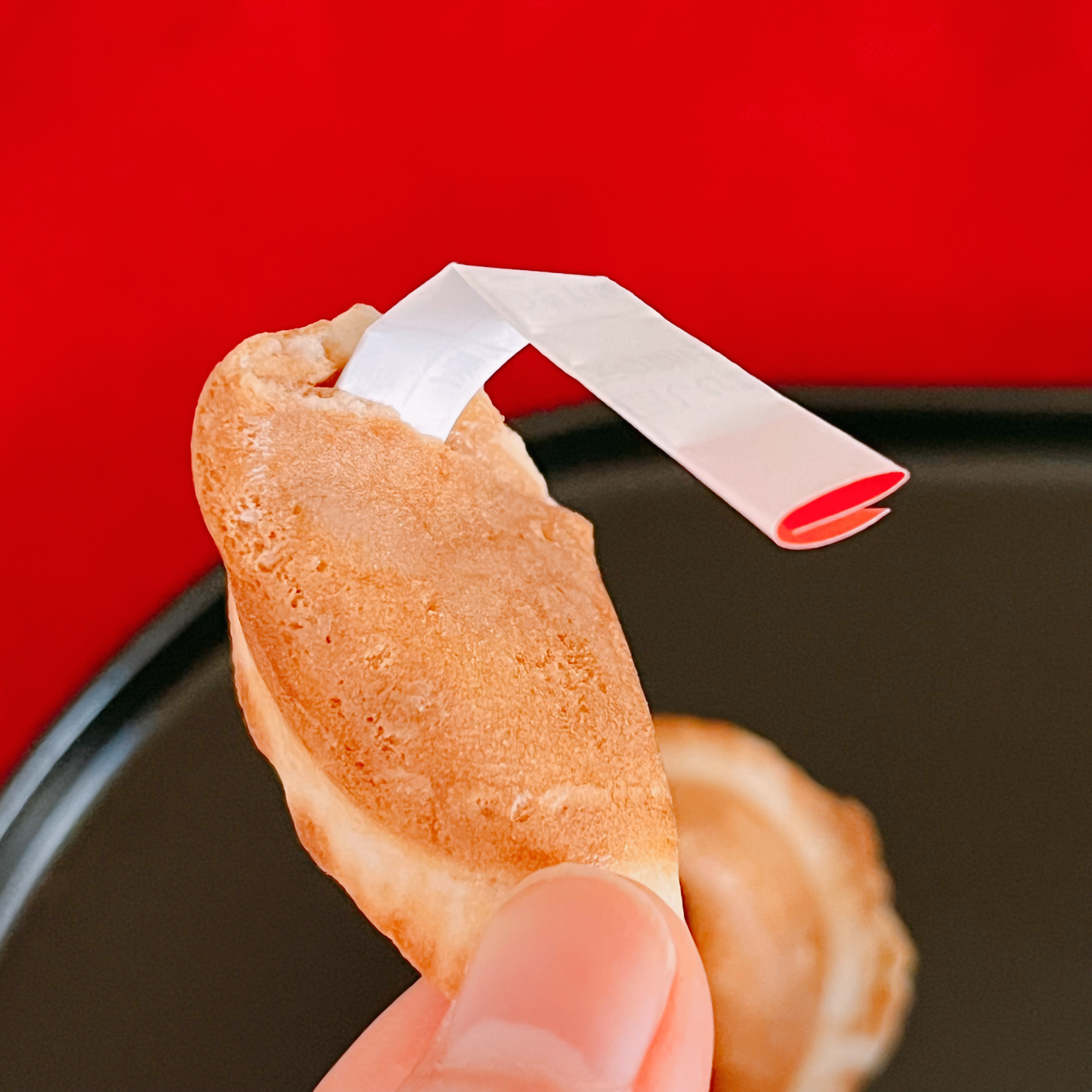 占いクッキーを持った手。クッキーからおみくじが少し出ている。背景に赤い布がある。