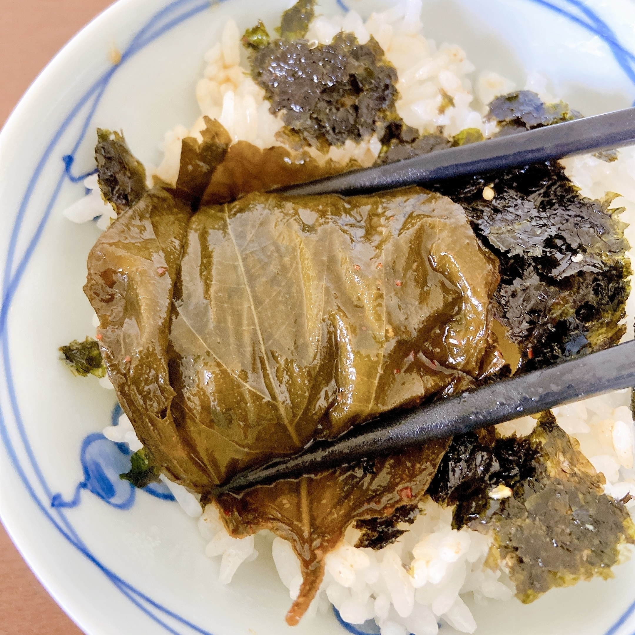 海苔と葉が乗ったご飯を箸で持ち上げている様子。料理の詳細が不明。