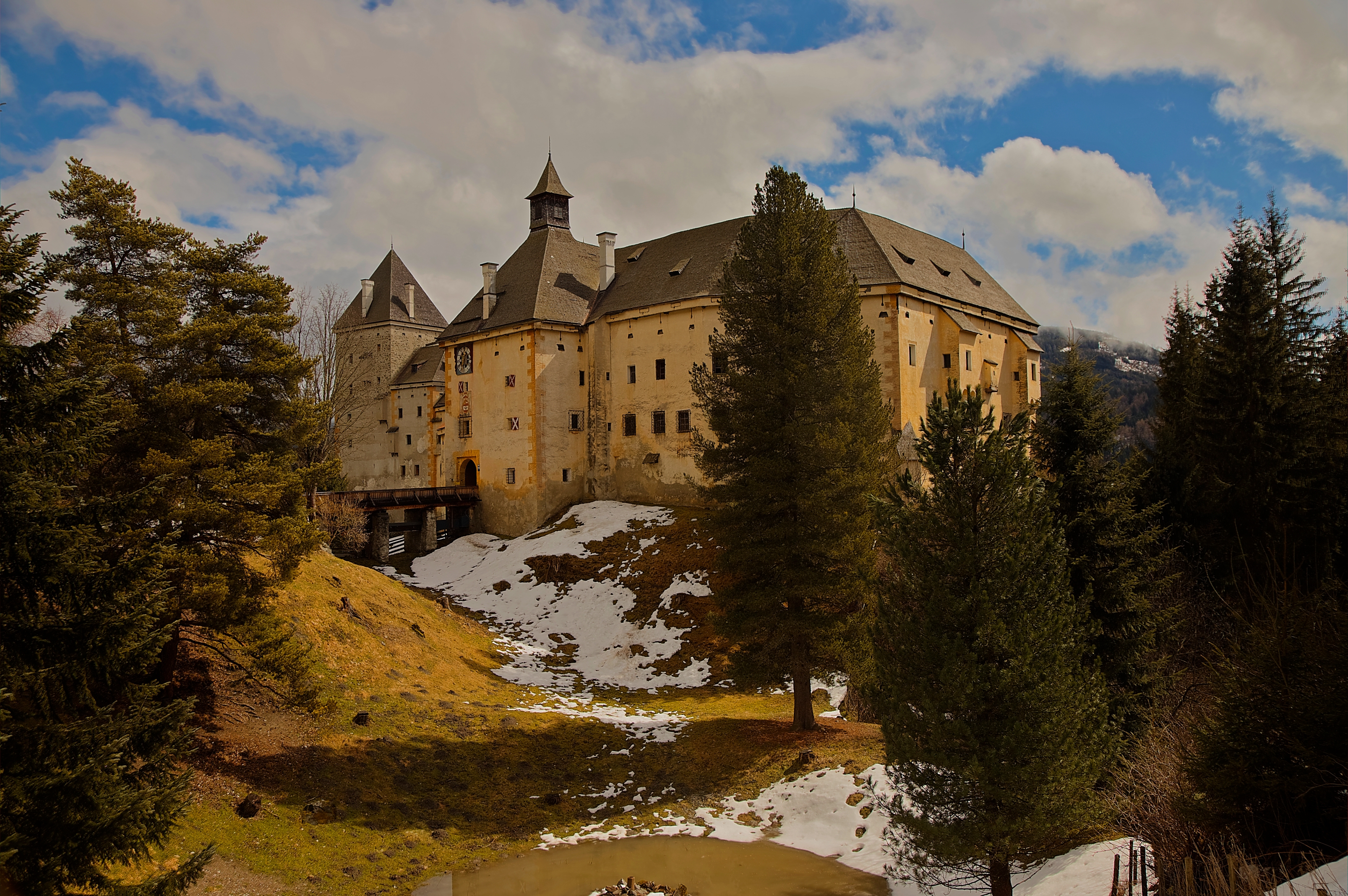 A historic castle with towers and conical roofs surrounded by trees and partially snow-covered landscape