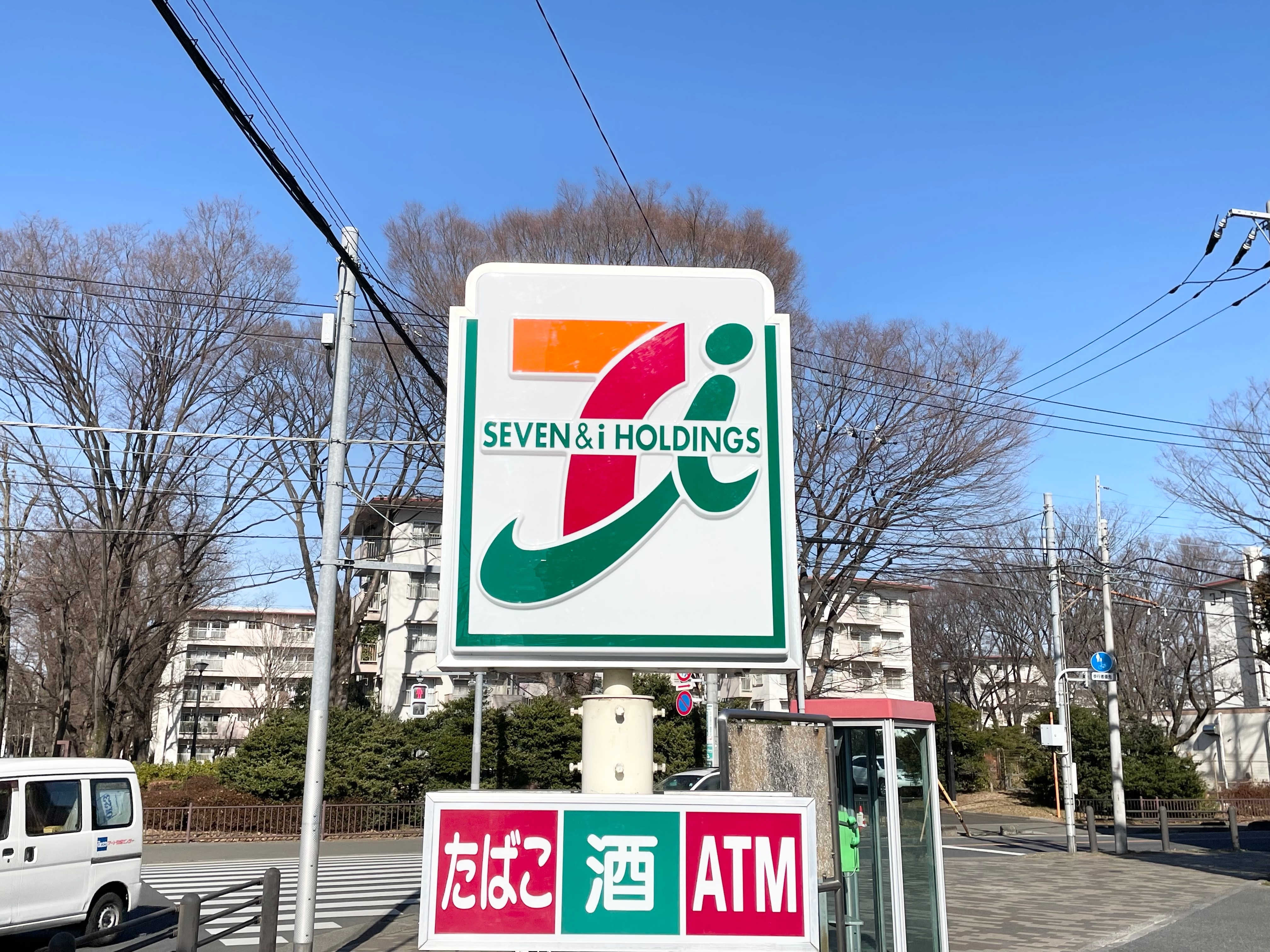 コンビニエンスストアの看板に「SEVEN & i HOLDINGS」と書かれたロゴがあり、下に「たばこ」「酒」「ATM」と表示。