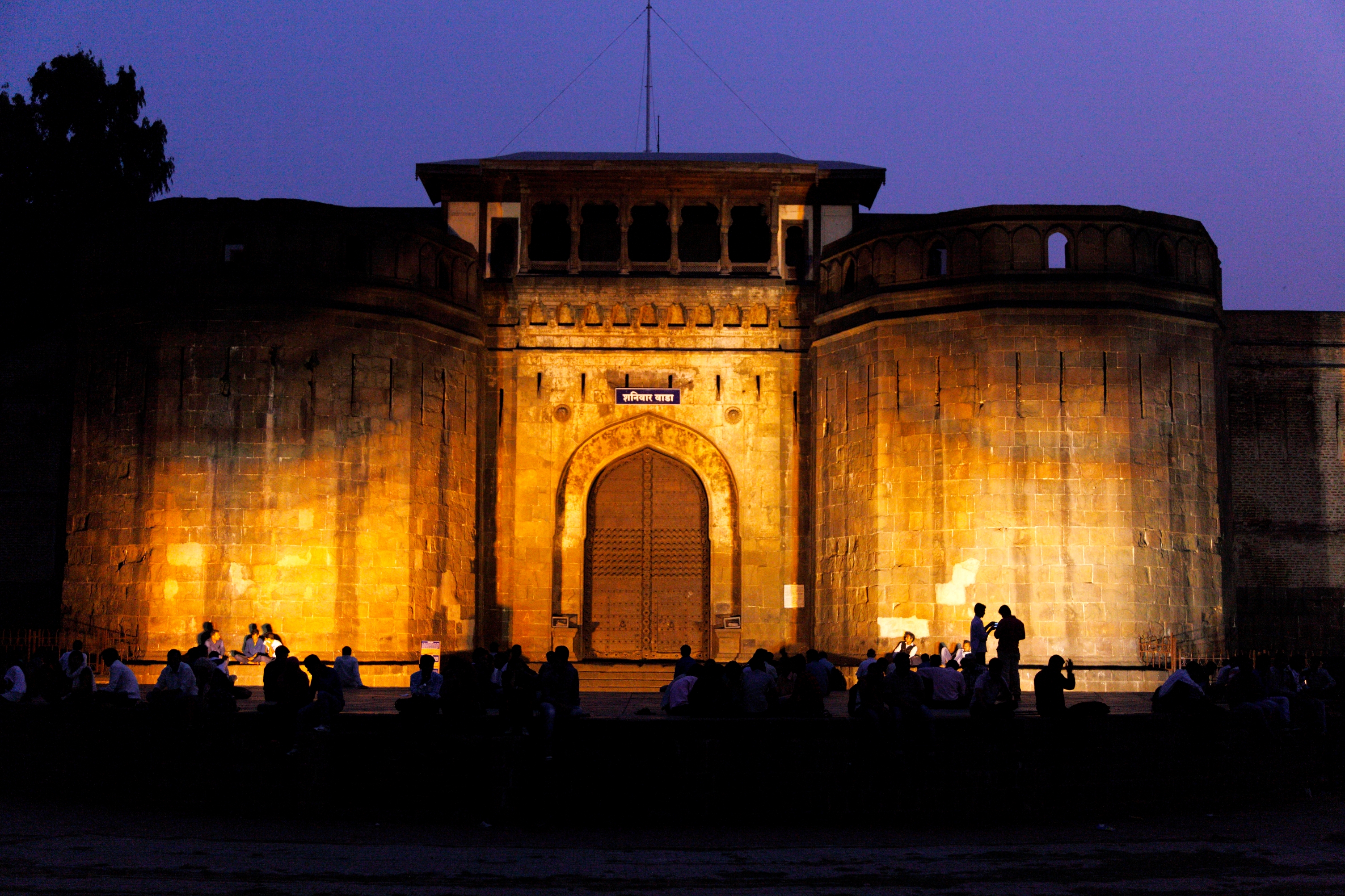 Large, illuminated historic fortress with arched entrance. Silhouetted people gather around the structure in the evening