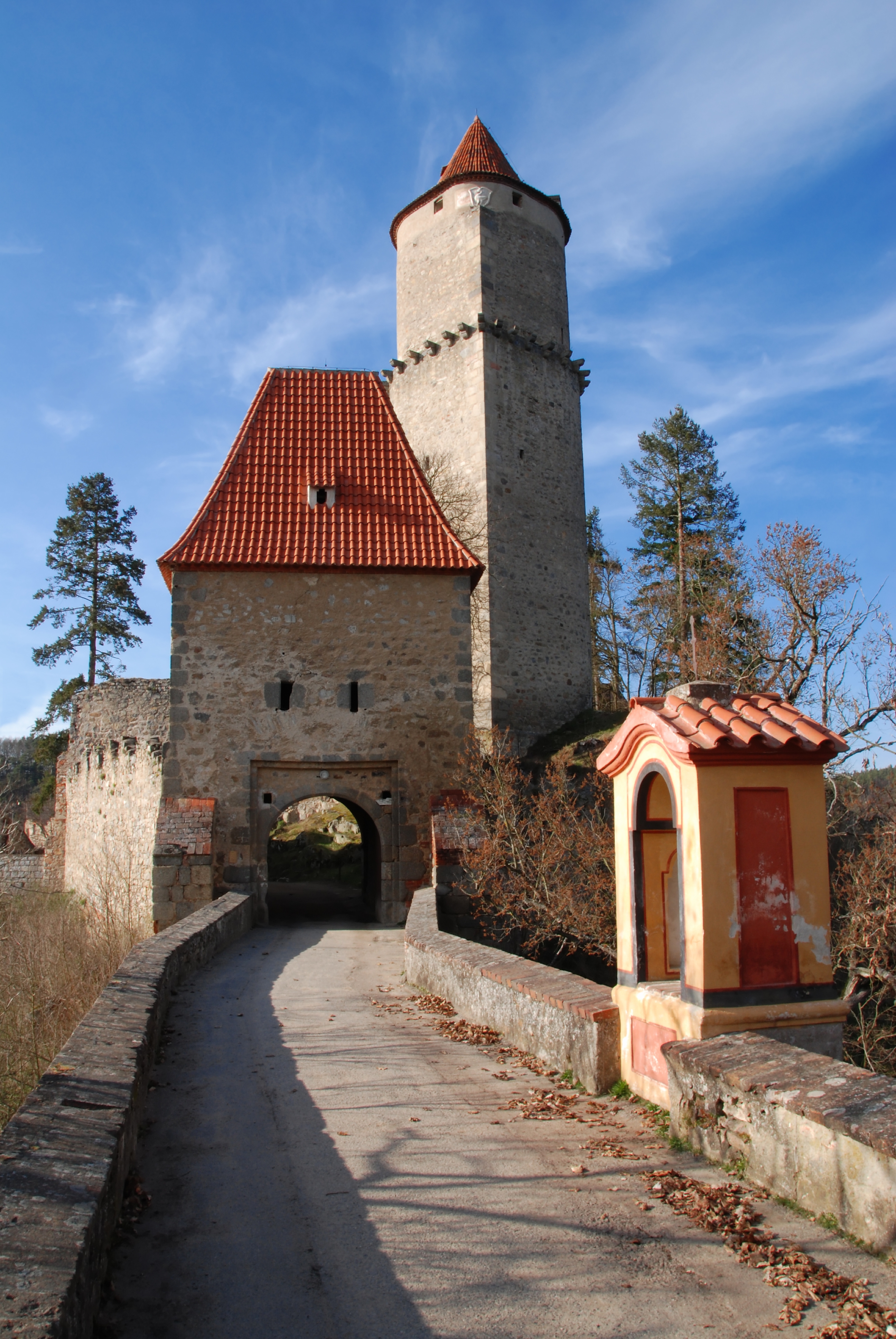 A medieval stone gatehouse with a tall tower and red-tiled roof, surrounded by trees and a winding path leading up to it
