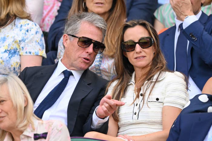 Two individuals seated at an event, wearing sunglasses and formal attire, engage in conversation