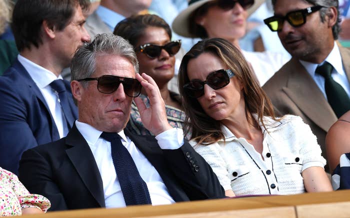 A man in a suit and tie sits with a woman in a casual outfit wearing sunglasses, surrounded by other people, at an outdoor event