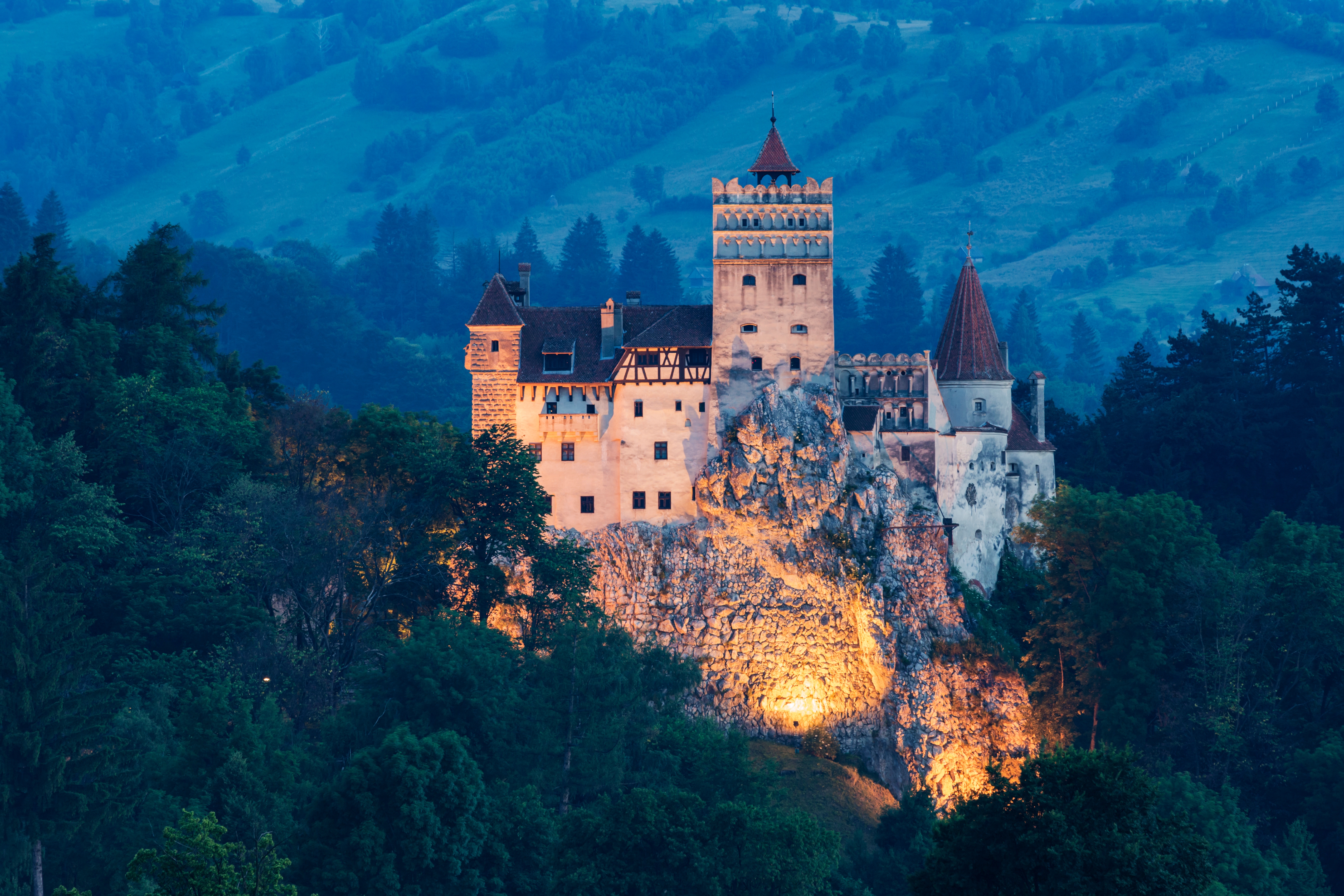 Castle surrounded by trees, atop a rocky hill, lit up at dusk with a mountainous backdrop