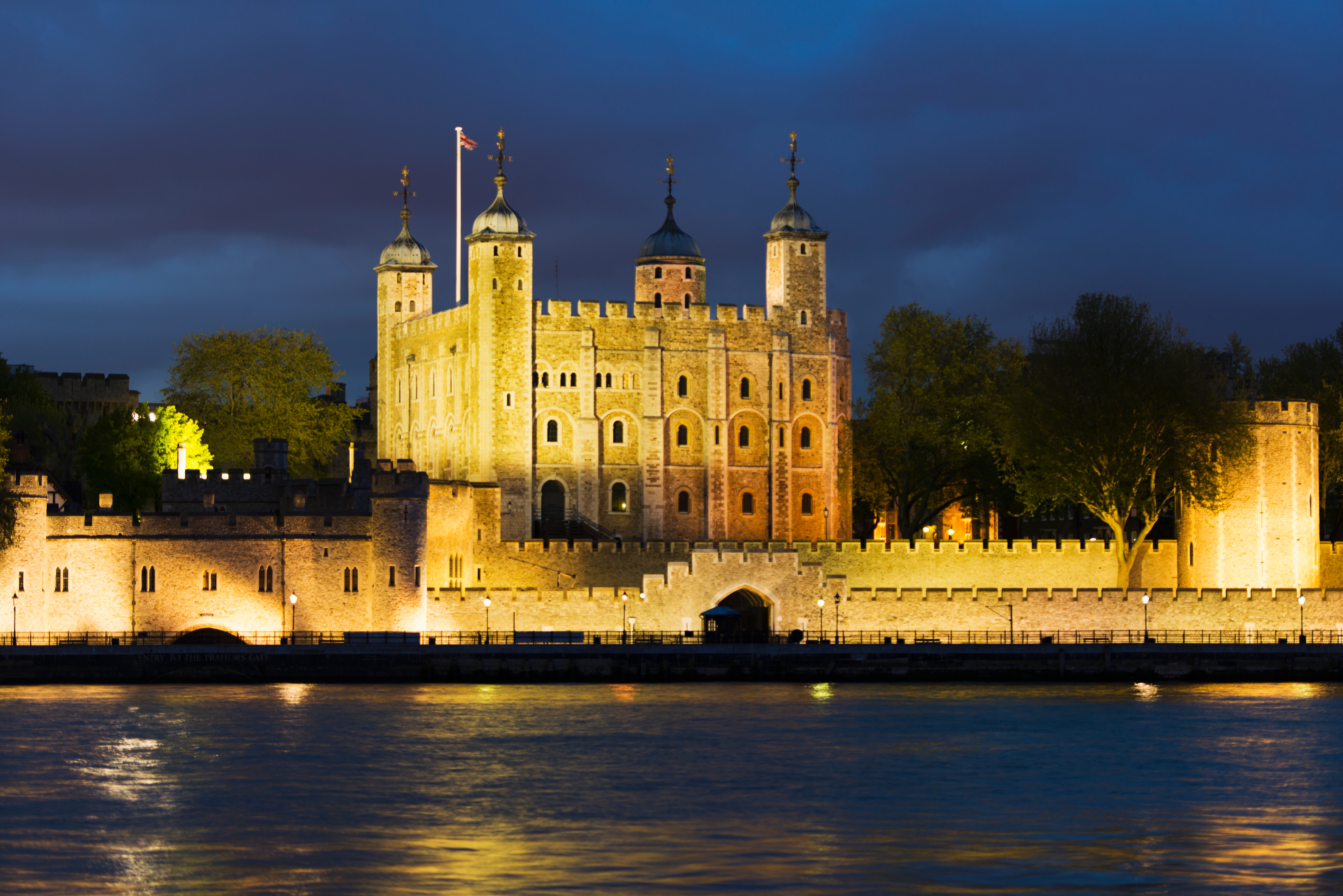 The image shows the Tower of London lit up at night, with the Thames River in the foreground