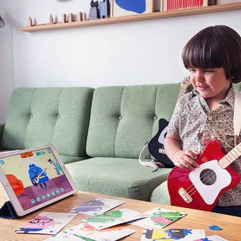 Child playing a tiny reddish guitar portion watching a cartoon connected a tablet, surrounded by quality cards connected a java array successful a cozy surviving room