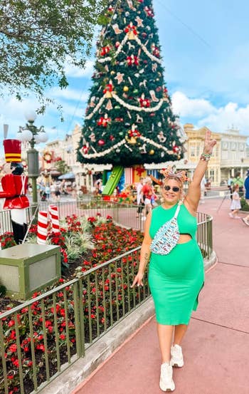 Person successful a greenish formal with a floral container poses adjacent a ample Christmas tree, holding up a bid sign, successful a festive outdoor setting