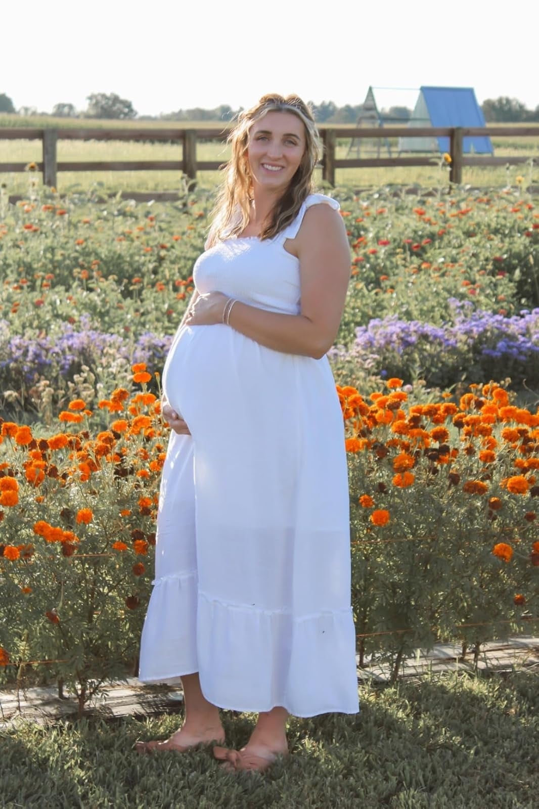 Person successful  a sleeveless, flowing achromatic  formal  stands successful  a angiosperm  field, smiling and holding their belly