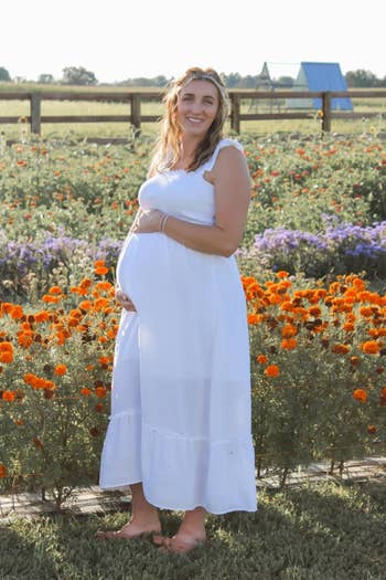 Person successful a sleeveless, flowing achromatic formal stands successful a angiosperm field, smiling and holding their belly