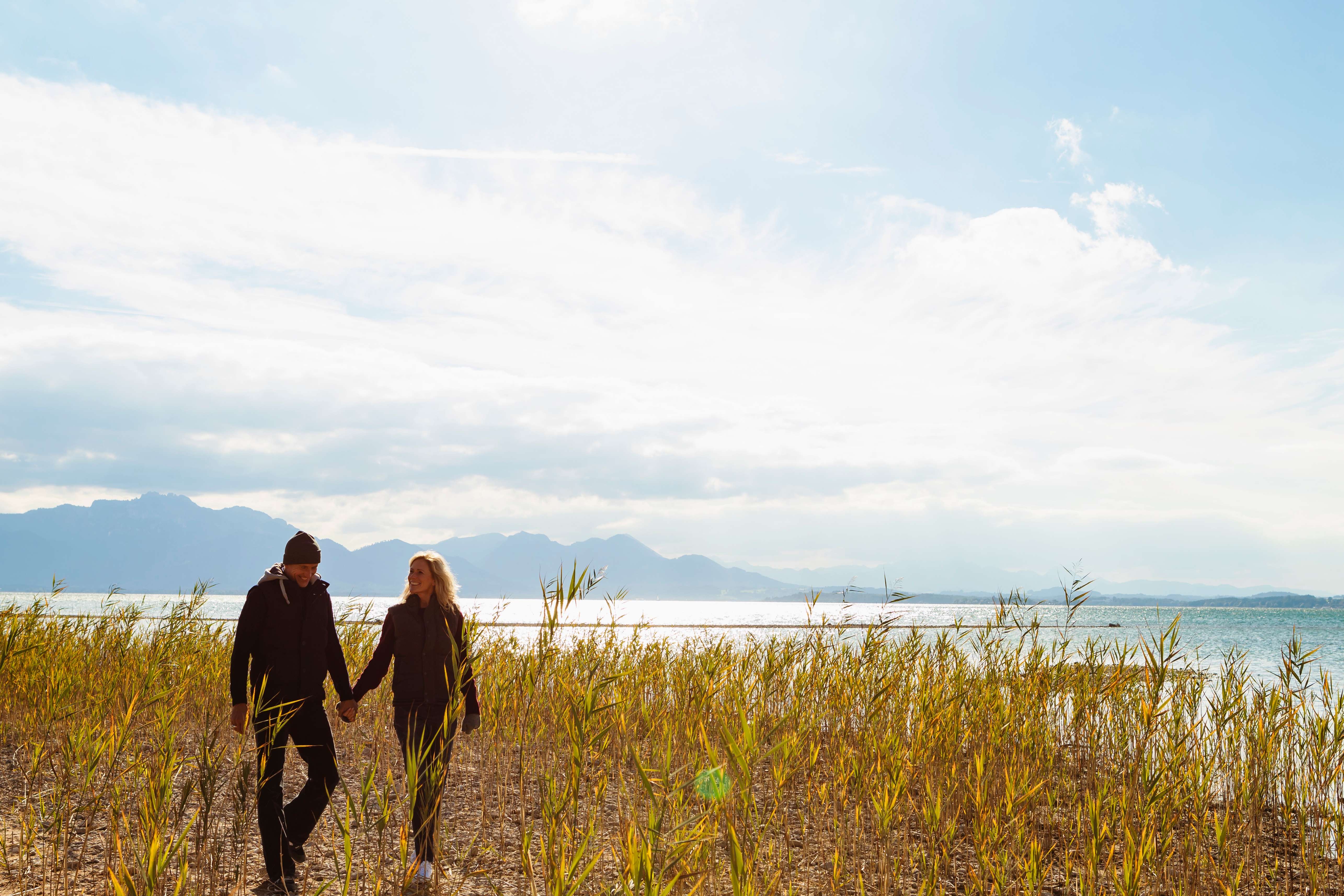 Et par som holder hender går langs en naturskjønn innsjø med fjell i det fjerne, under en lys himmel
