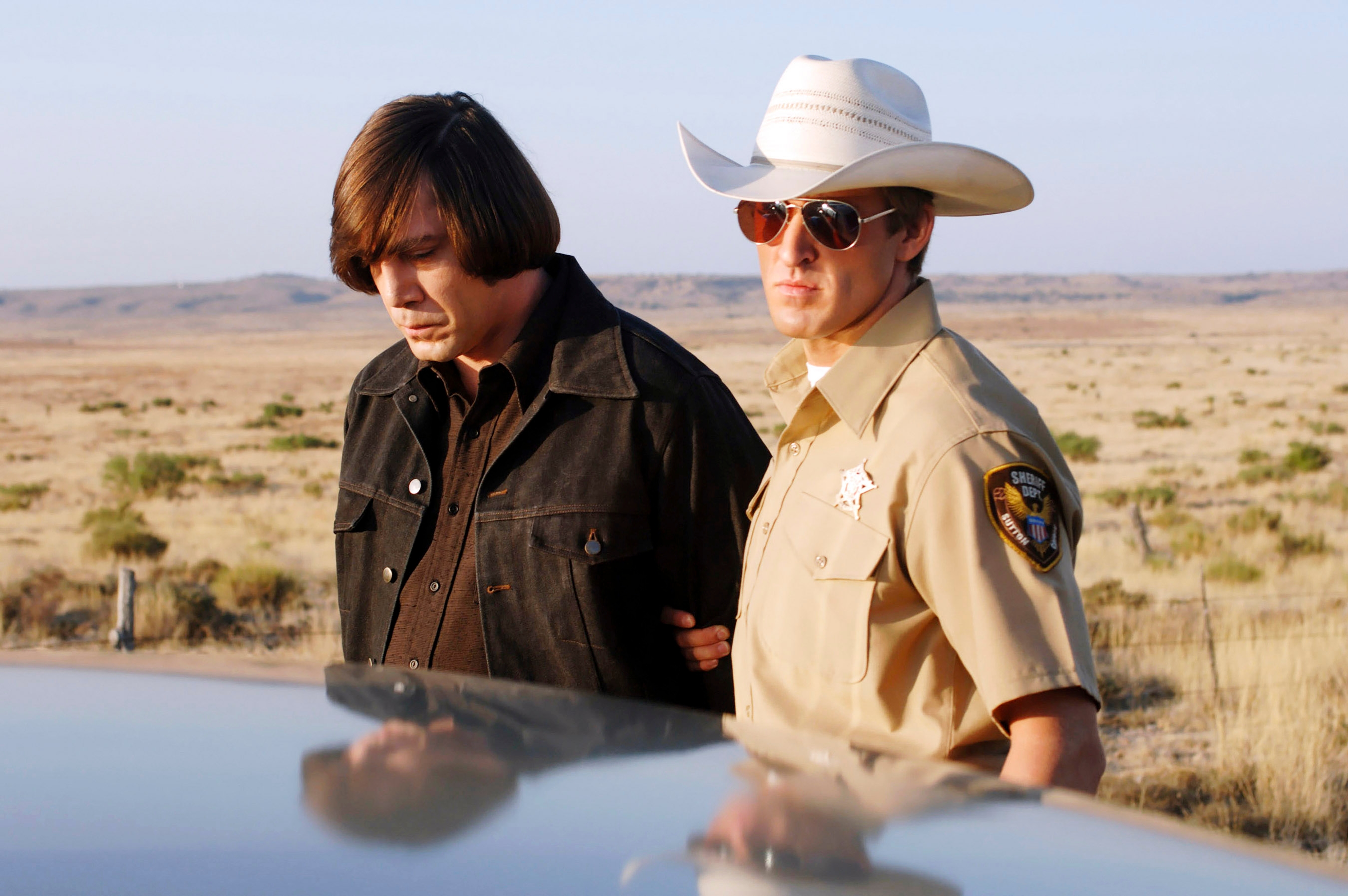 A scene from a film with a man in a leather jacket being handcuffed by a sheriff in a cowboy hat and sunglasses in a desert landscape