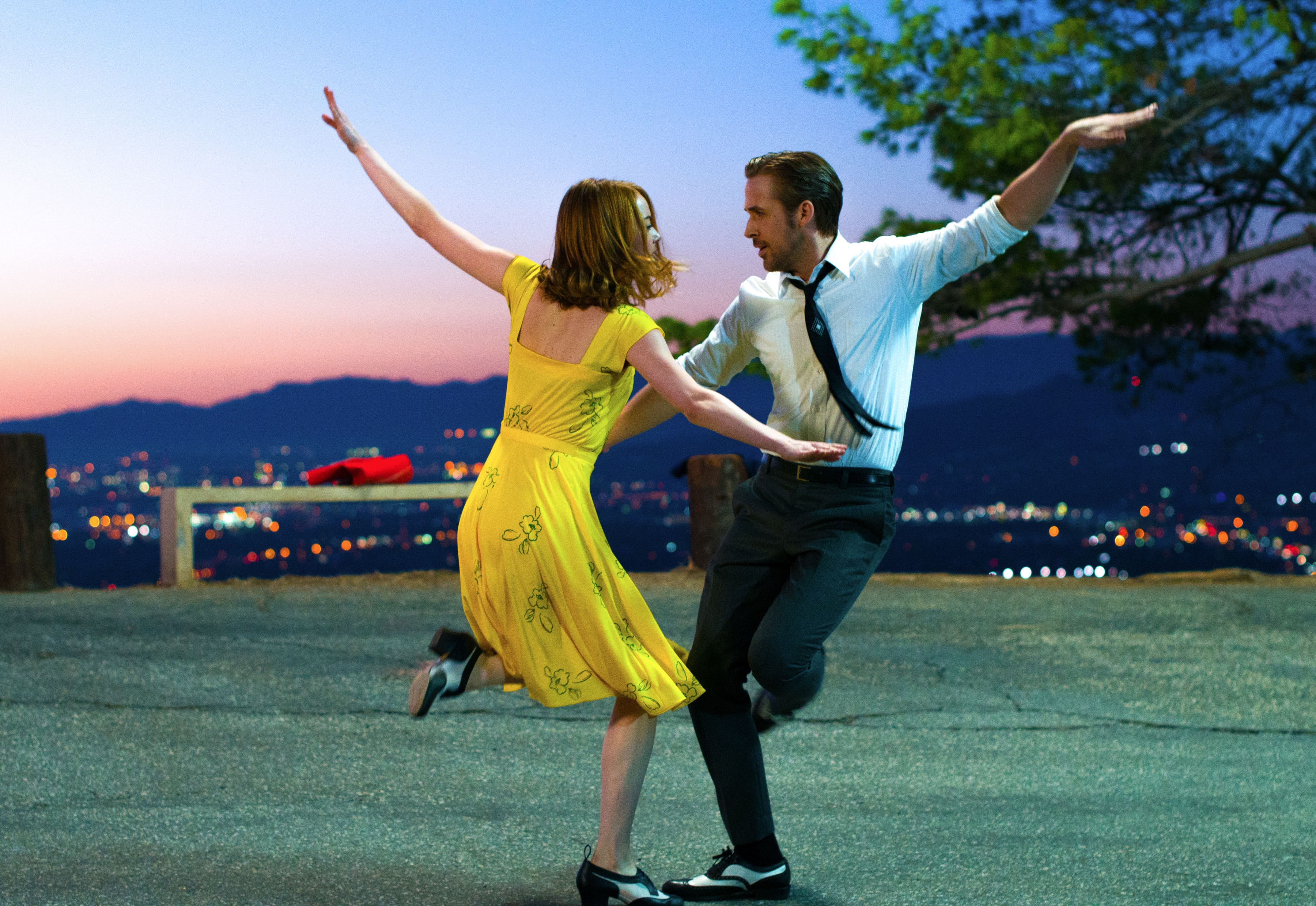 Two people joyfully dance on a hilltop at sunset, one in a bright dress and the other in a shirt and tie. City lights twinkle in the background