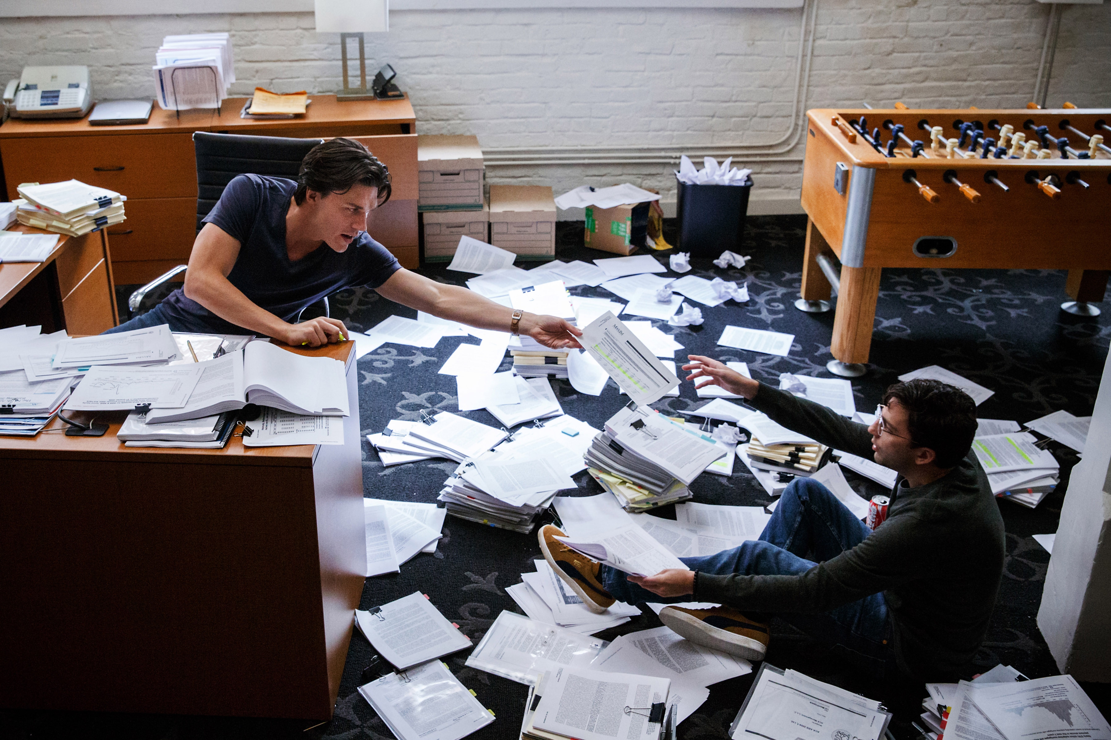 Two people surrounded by scattered papers in an office; one is seated on the floor, extending a document towards the other, who is bending over a desk