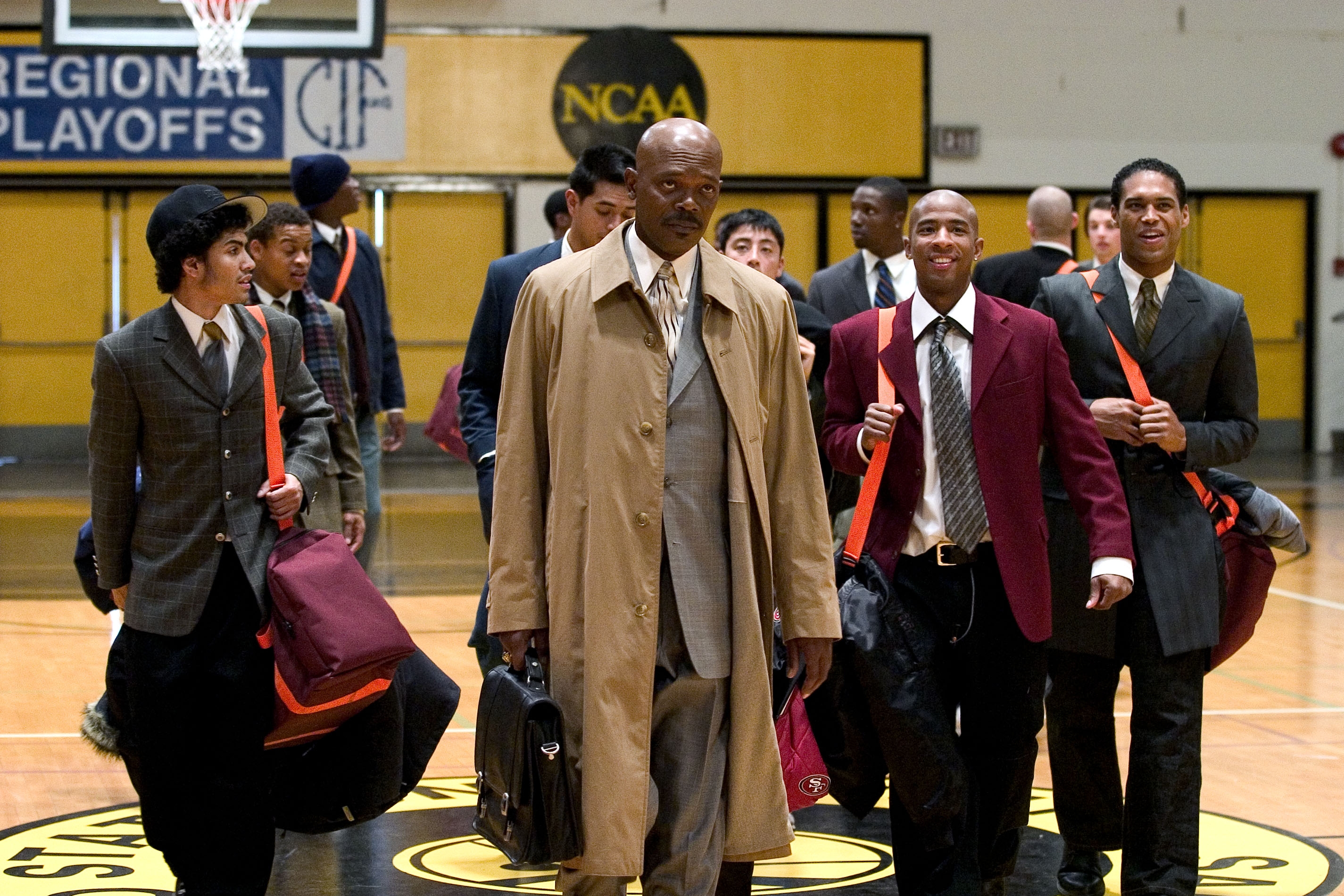 A group of well-dressed men walk confidently on a basketball court. The central figure wears a long coat
