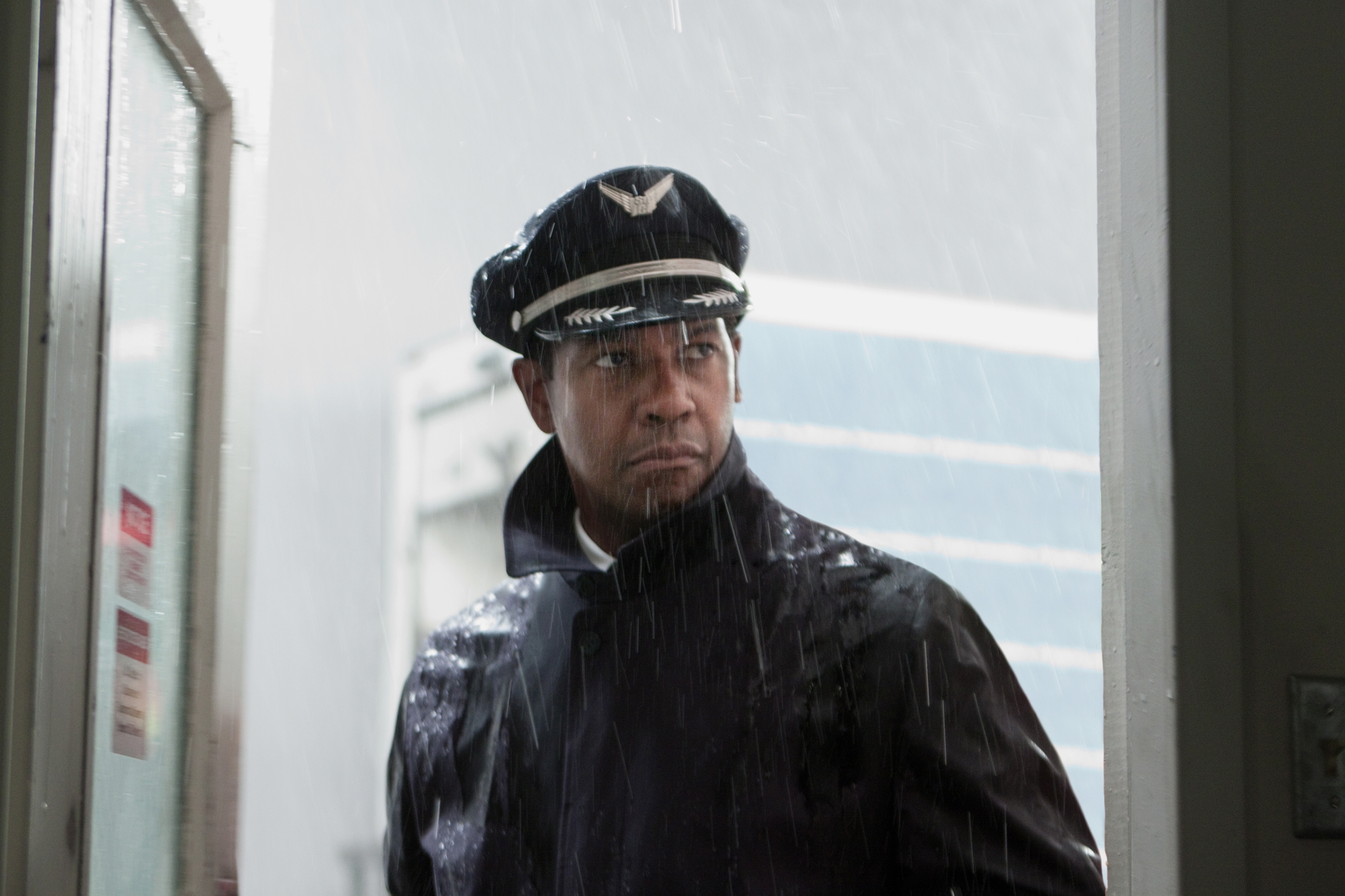 Man in pilot uniform stands in doorway, looking outside with a serious expression, heavy rain pouring down