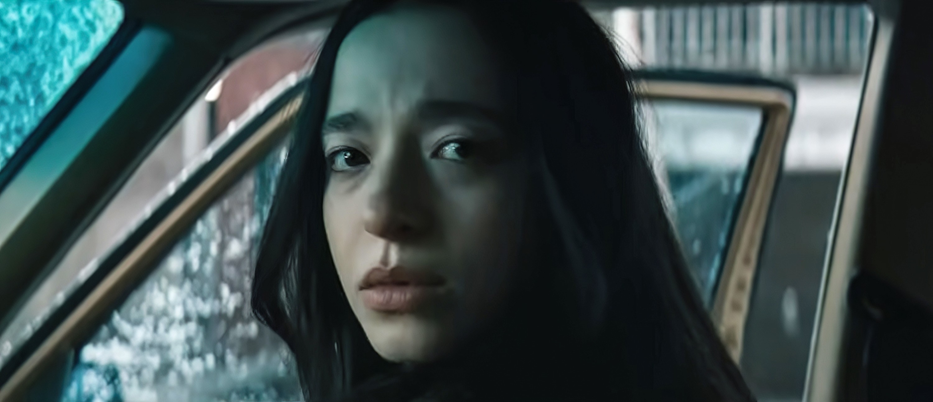 Person with long hair sits in a car, looking pensively out of an open door. Snow is visible outside