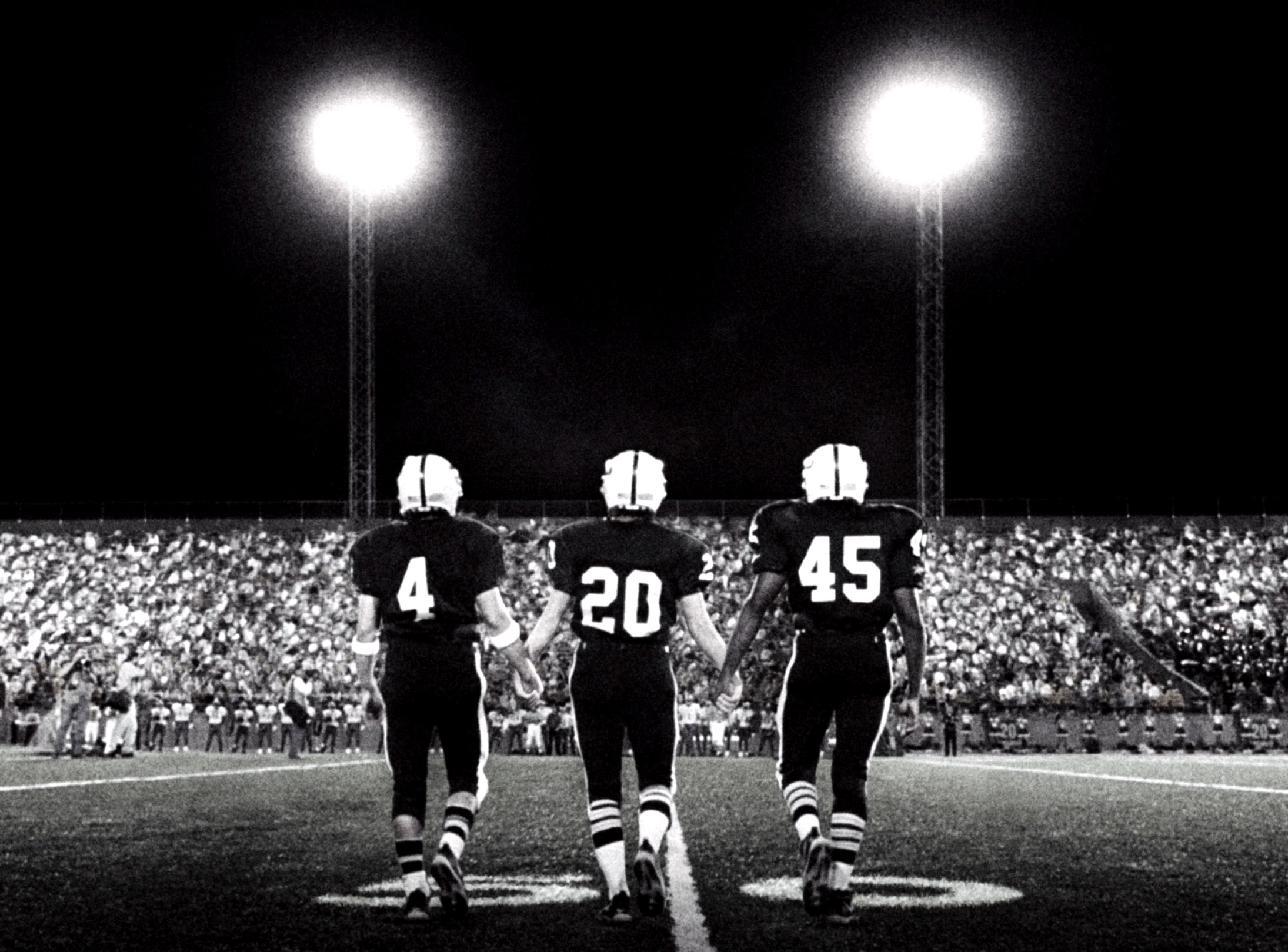 Three football players walk onto a lit field at night, facing a large crowd. They wear helmets and numbered jerseys: 4, 20, and 45