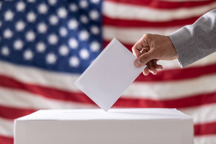 A hand in a suit places a ballot into a box in front of a blurred American flag, symbolizing voting or election participation
