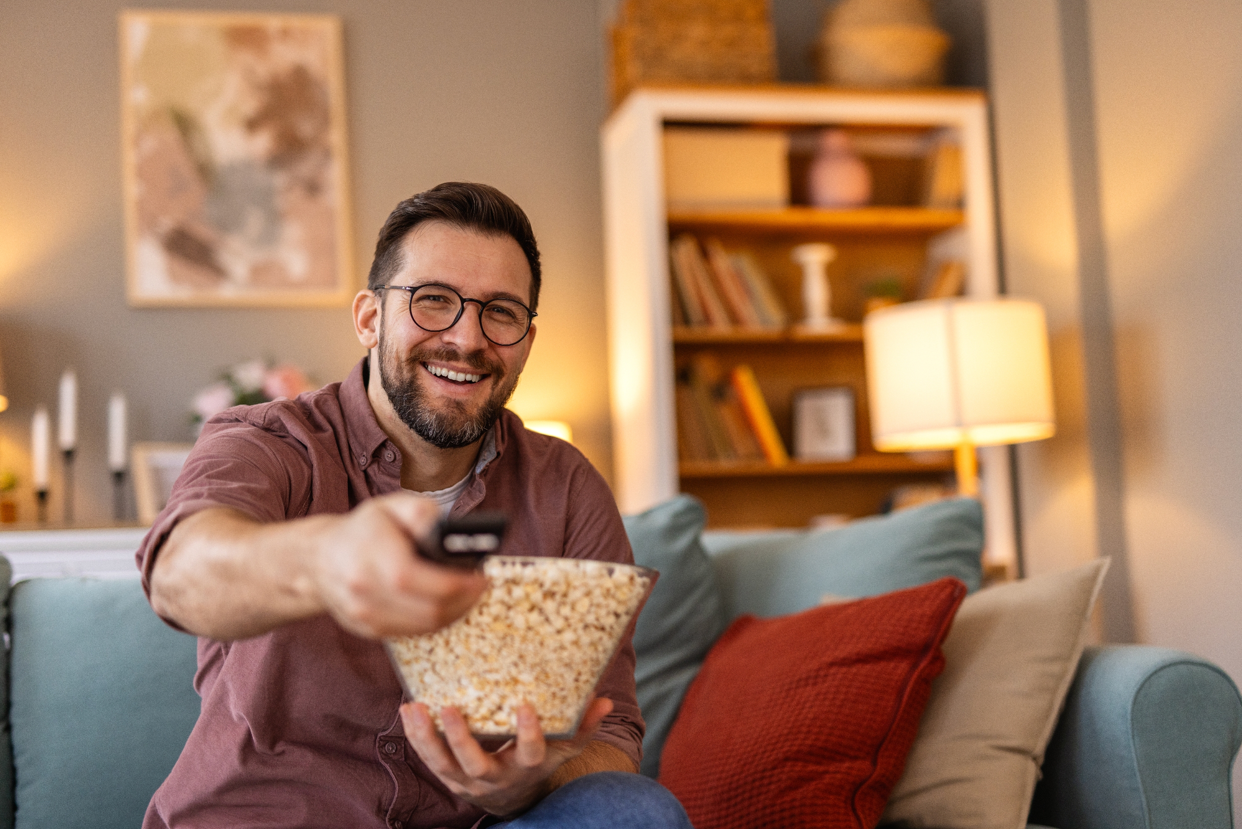 Man with glasses smiling, sitting on a couch holding a remote and a bowl of popcorn in a cozy living room setting