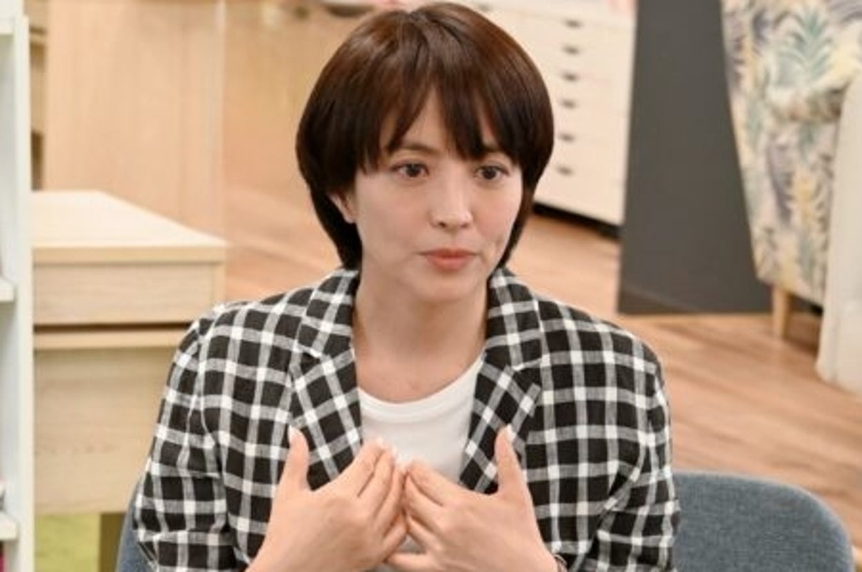 女性がチェック柄のジャケットを着て椅子に座り、別の人物と話している。背景に本棚とソファがある。