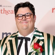 Person on red carpet in a green and white striped suit with a red rose boutonniere, wearing glasses and a bow tie, smiling at the camera