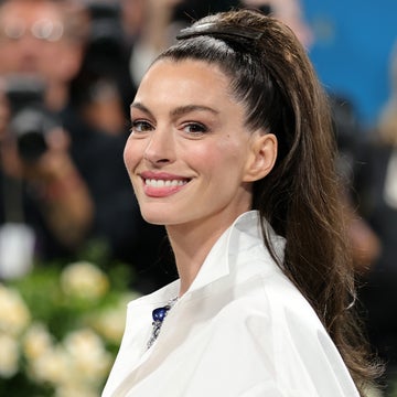 A person smiles on a red carpet, wearing a white shirt with puffed shoulders. Photographers and floral decorations are in the background