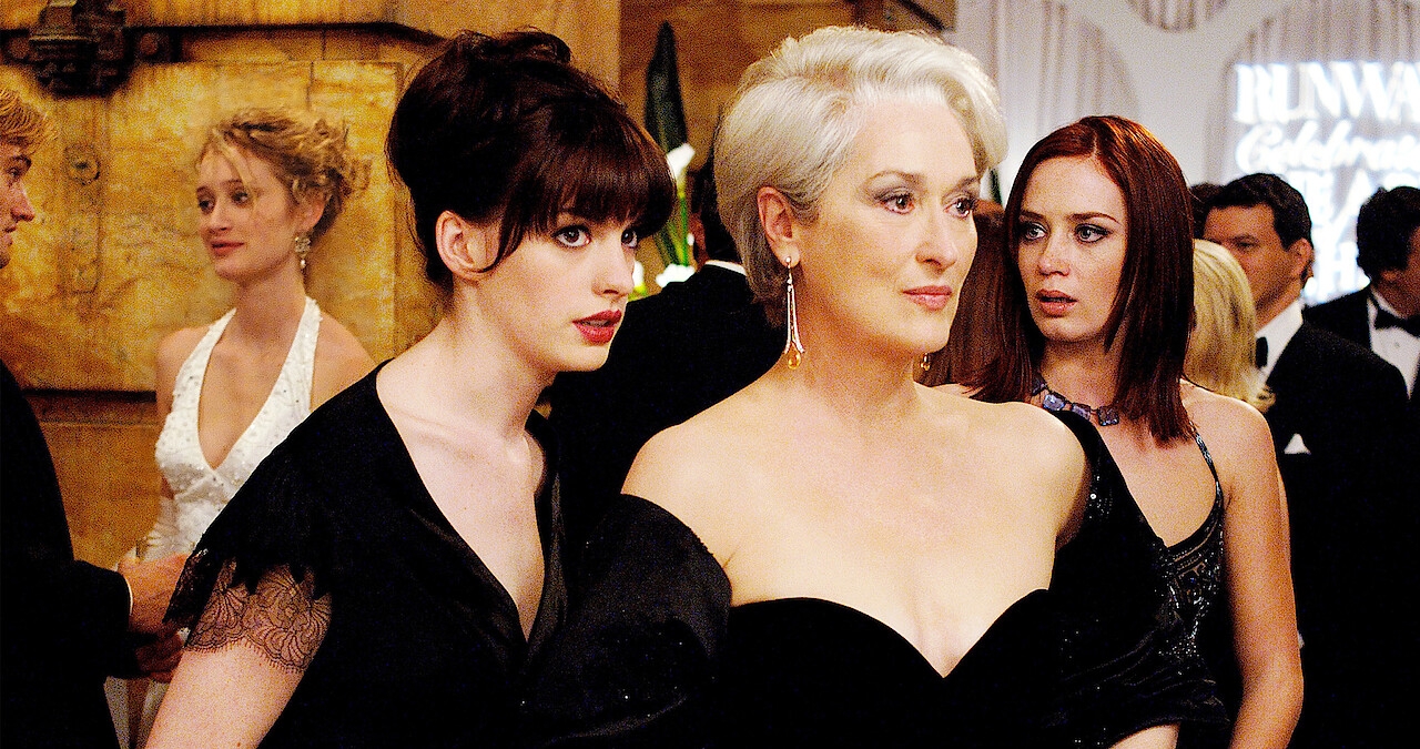 A scene from a movie with a woman in an elegant strapless gown beside another in a chic black dress, surrounded by formally dressed people