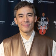 Person smiling on theatre awards red carpet, wearing a leather vest over a collared shirt. Awards backdrop with logos visible