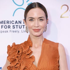 Person in pleated dress with a large ruffled shoulder detail at an event, posing in front of a backdrop with logos and text