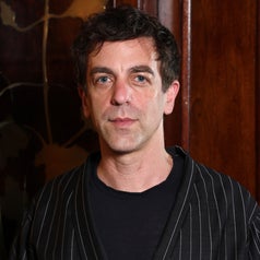 Person wearing a dark pinstripe suit jacket over a black shirt, standing in front of a wooden backdrop
