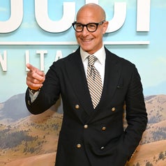 Person in double-breasted suit with glasses smiles and gestures in front of a scenic backdrop at an event