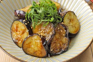 焼きナスの薄切りが器に盛られ、大葉とごまがトッピングされている。横に箸が置かれている。
