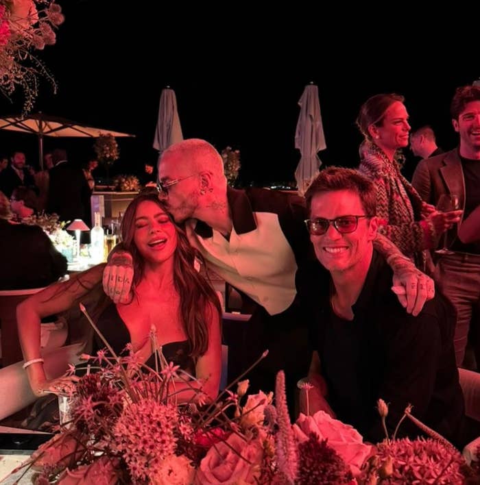 Three people pose at an upscale outdoor event, with one giving a kiss on the cheek. They are surrounded by floral decorations and warm lighting