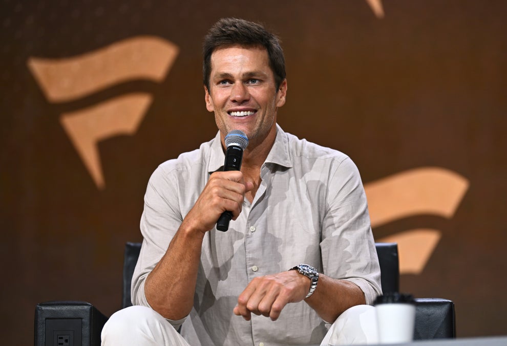 Person smiling, holding microphone, seated. Wearing casual light shirt and white pants