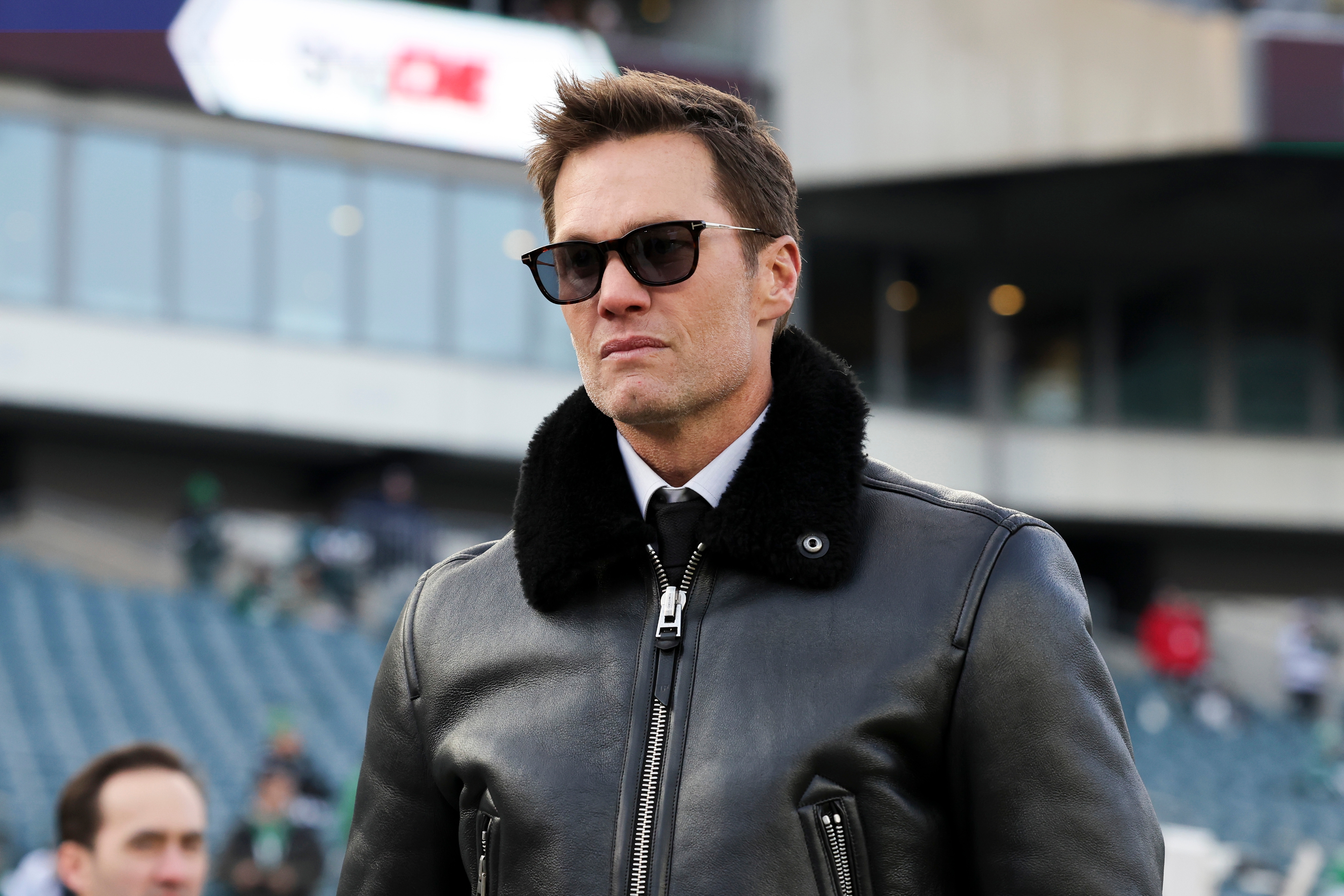 A person in sunglasses wearing a leather jacket, standing in a stadium setting