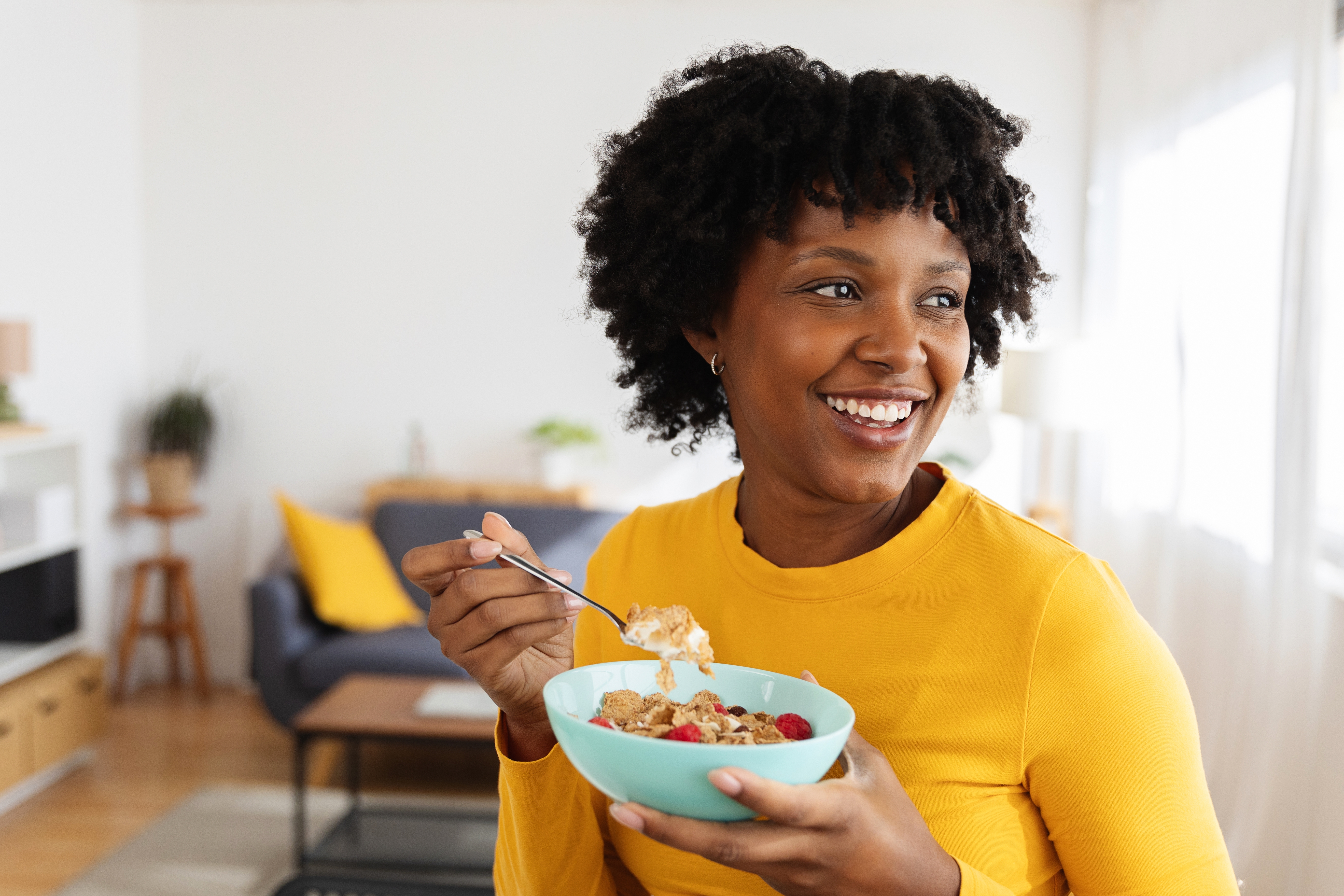 Person smiler mens du spiser frokostblandinger med frukt i en bolle, sitter i en koselig stue -omgivelser