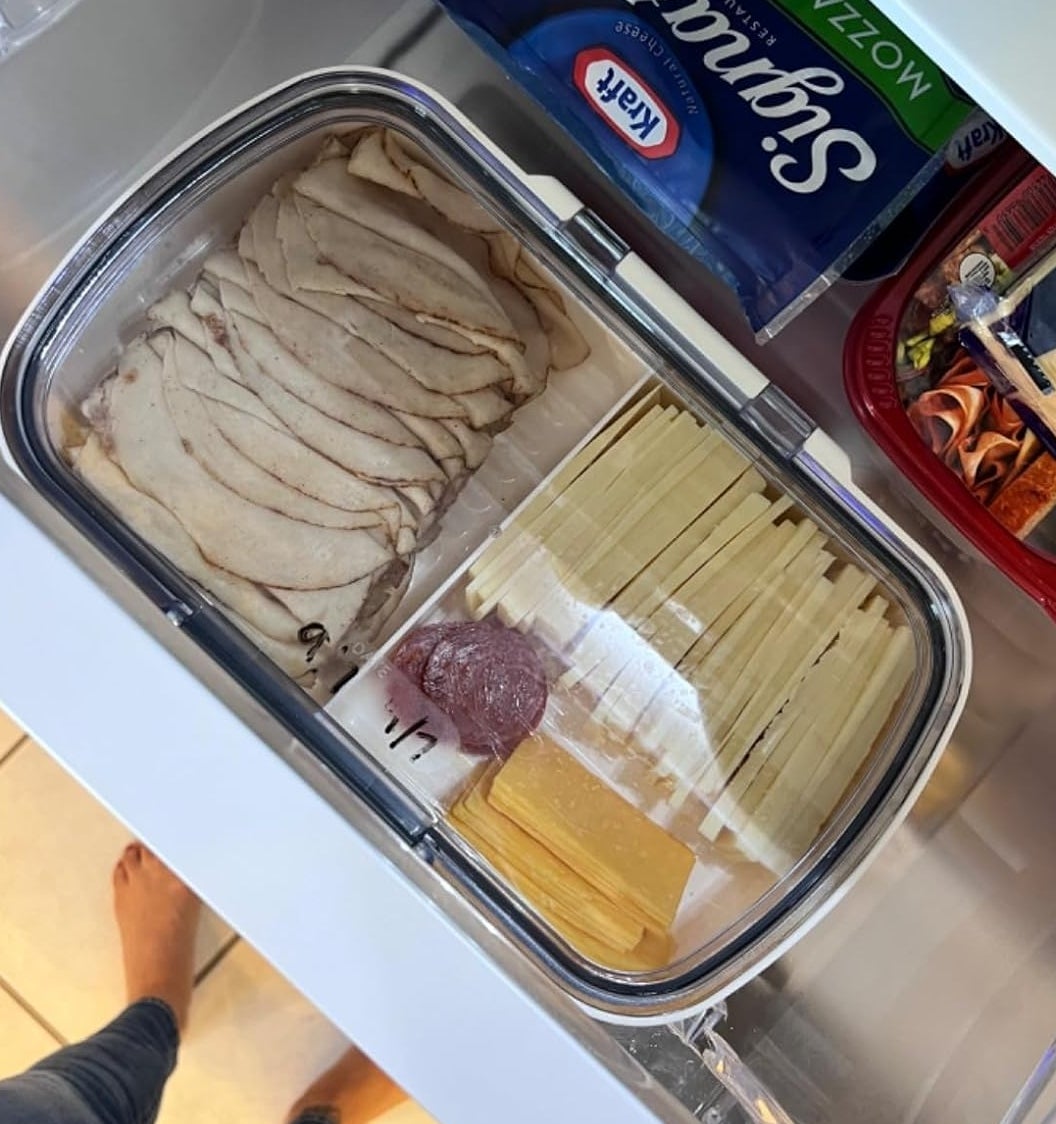Open fridge drawer with storage container holding deli turkey slices, cheese slices, and a small sausage. Mozzarella and other packaged items are visible